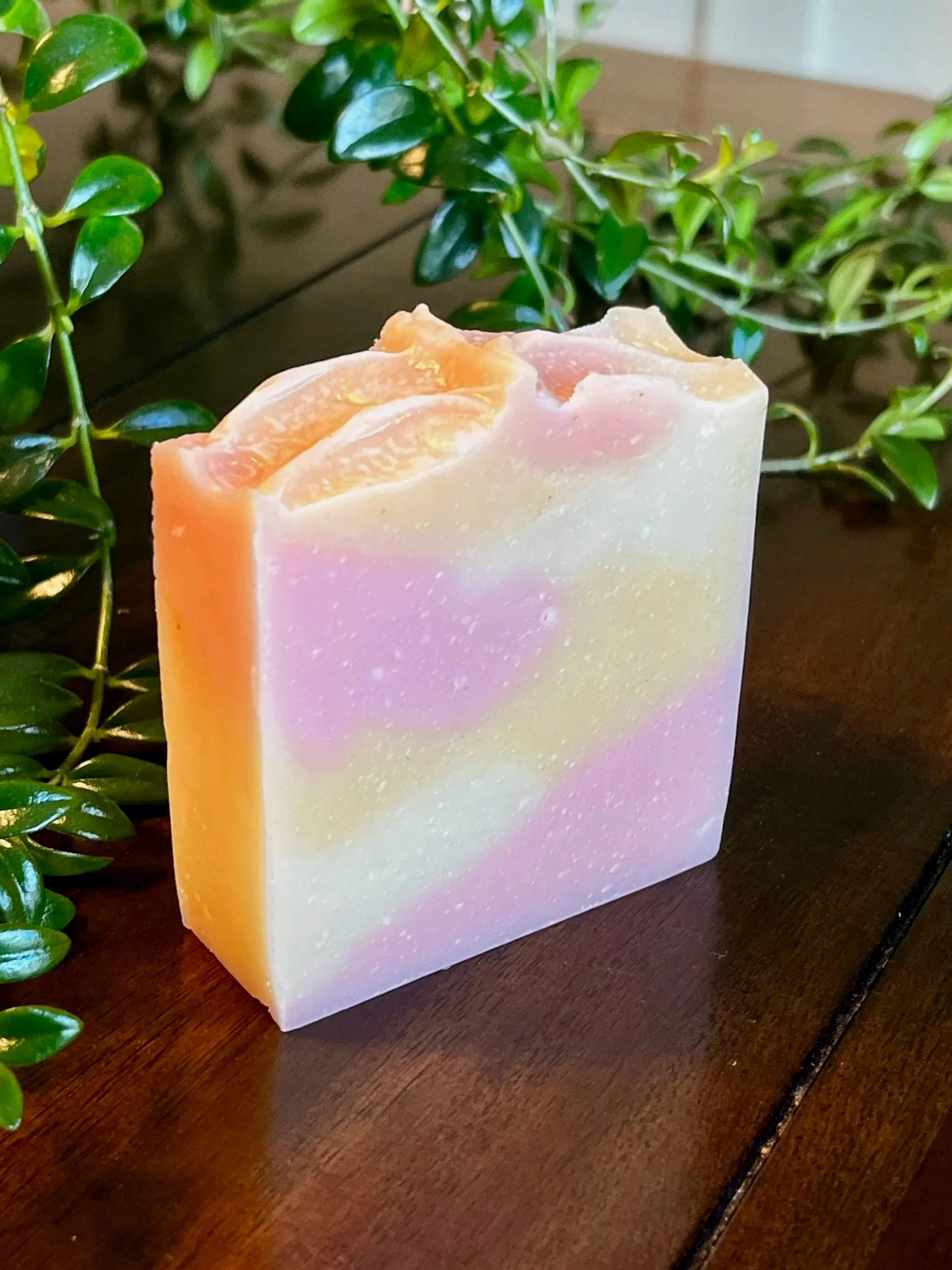 Colorful soap bar with pink, yellow, and white swirls placed on a wooden surface with green leafy plants in the background.