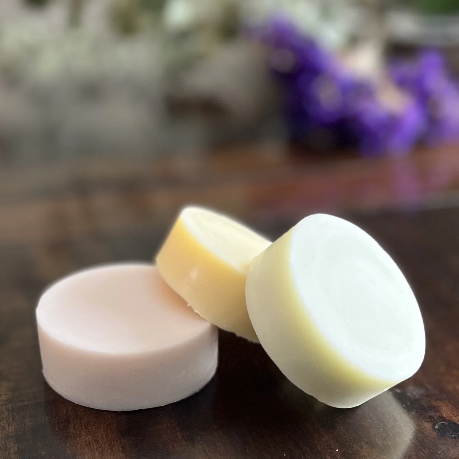 Close-up view of three sliced cheese pieces in various pastel colors on a dark wooden surface with a blurred background featuring purple flowers.