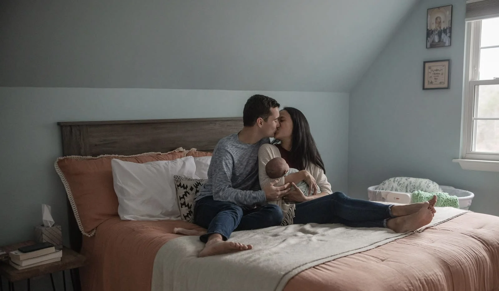 At-home newborn session in Harrisonburg, Virginia — lifestyle photography of parents cuddling baby on the bed in soft natural light.