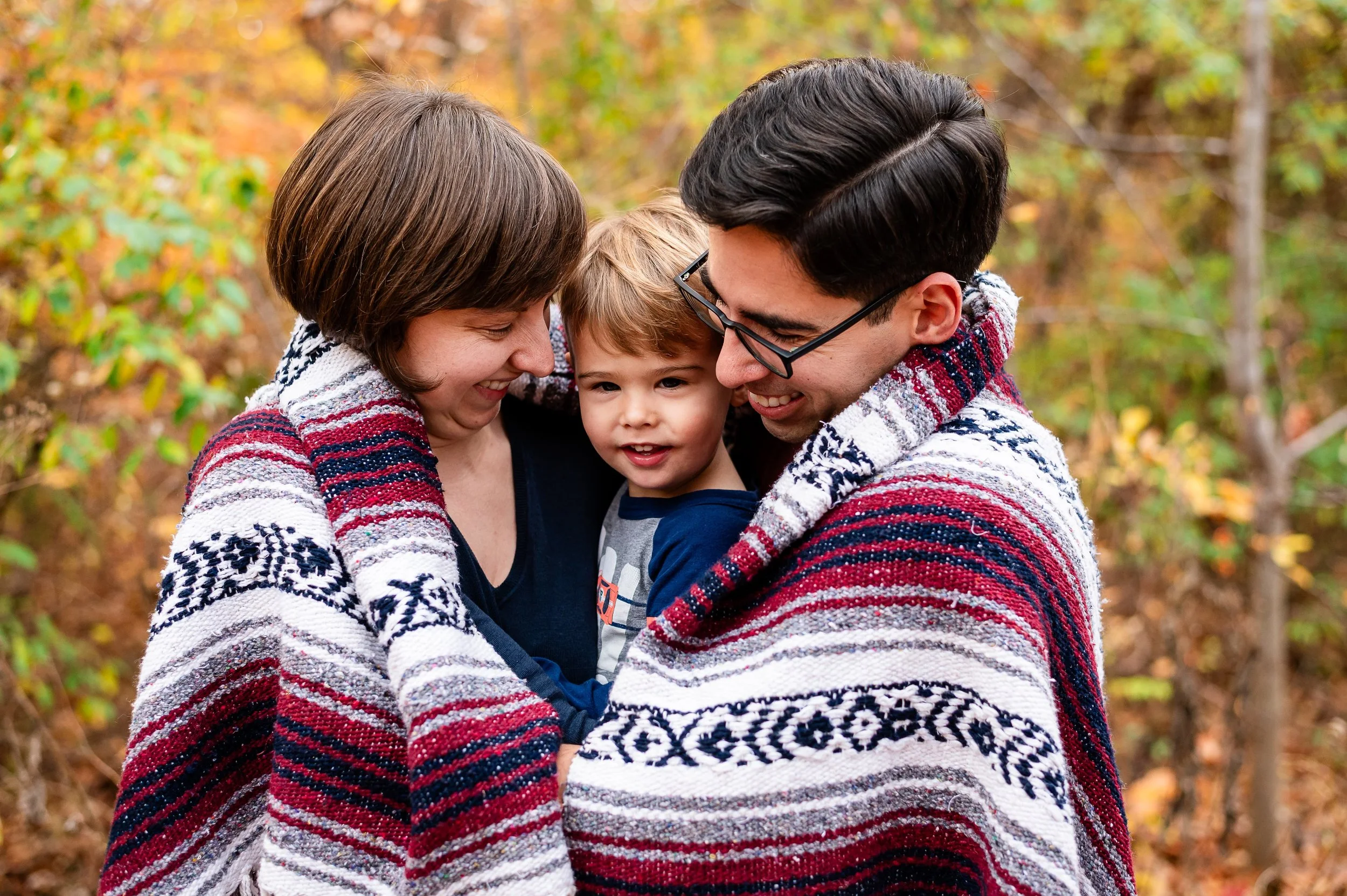 father and mother holding their toddler son while covered in a blanket during a fall mini session