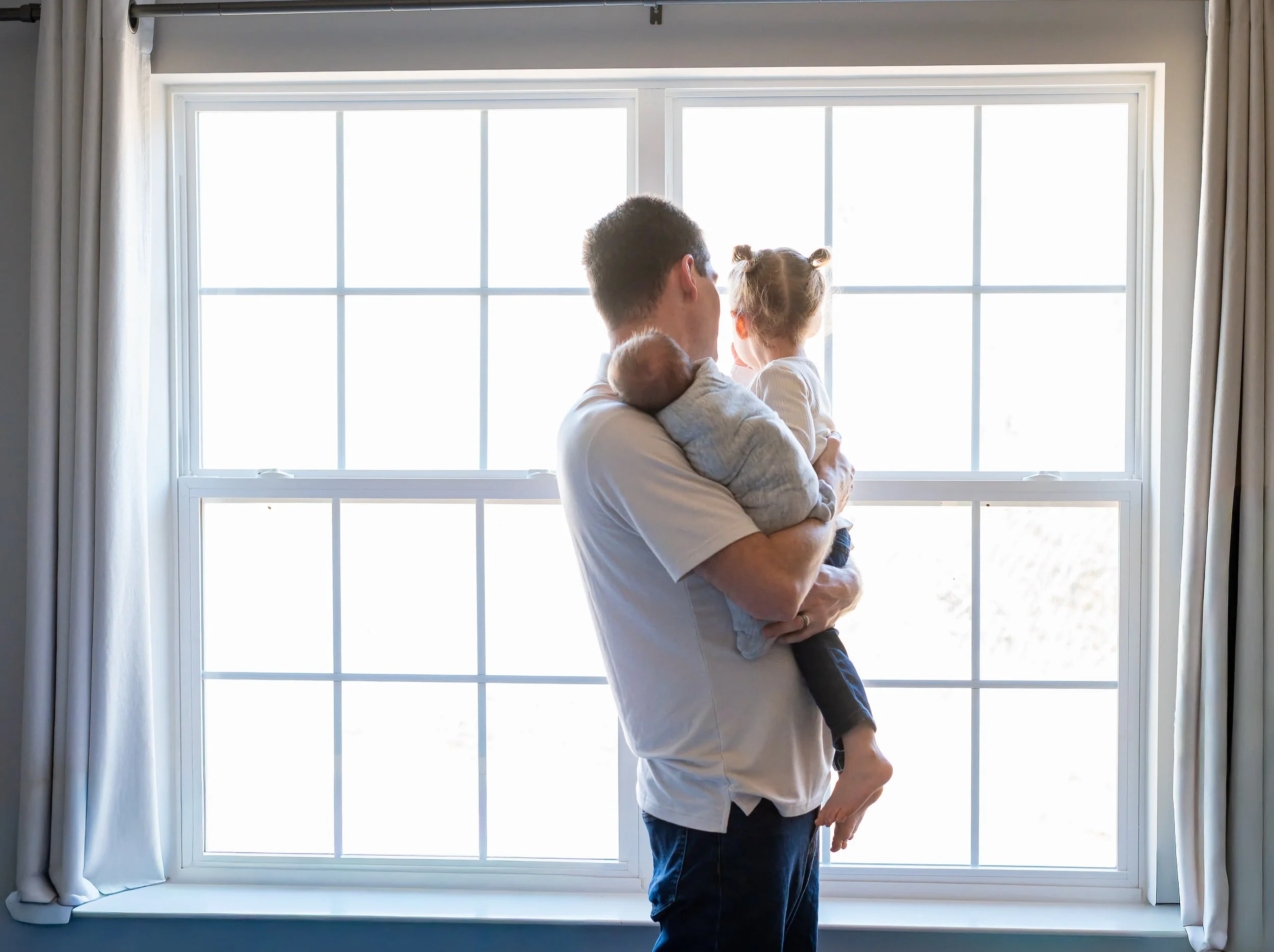 Parent holding a newborn and young child in front of a large window during an in-home family photography session.
