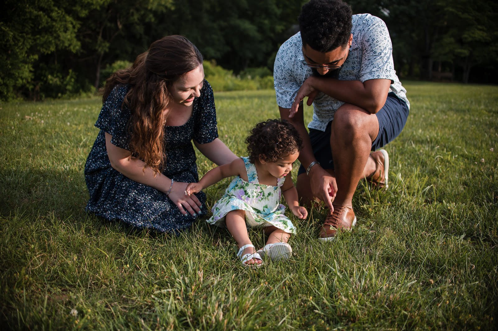 mom and dad sitting in the grass playing with toddler girl in a forest in Charlottesville, VA during a maternity session With Erin E Gallagher Photography