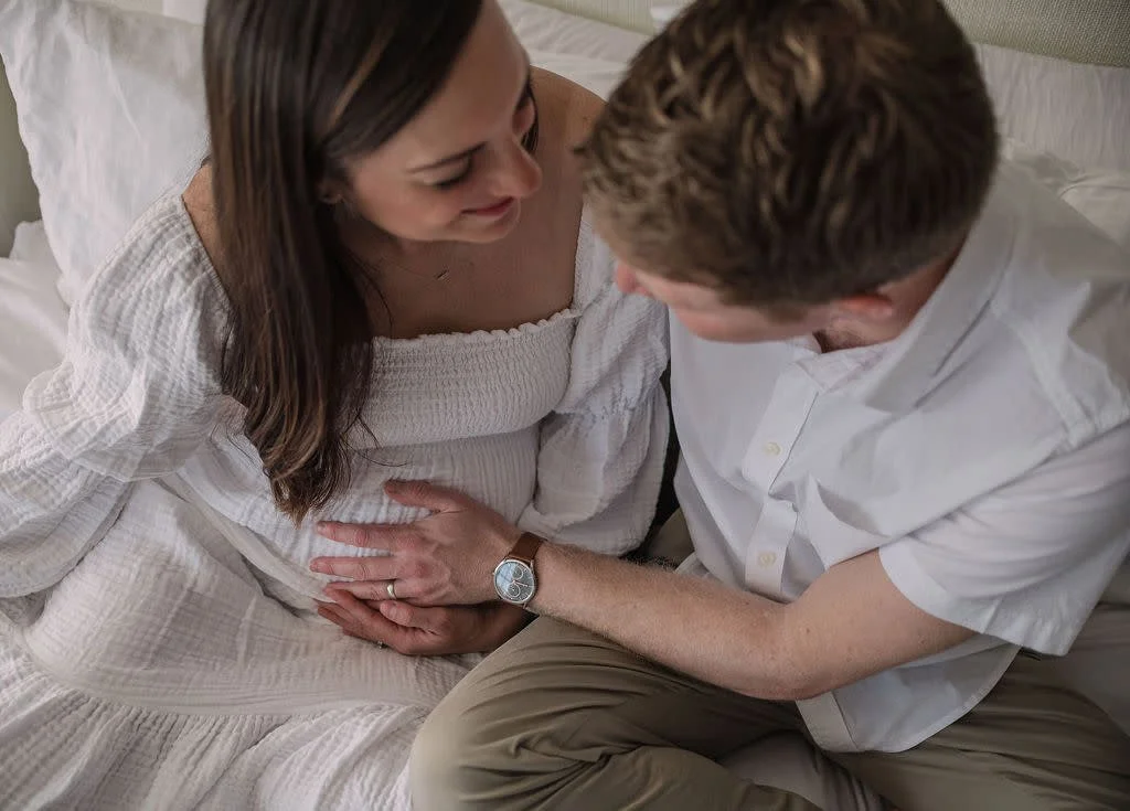 Husband and wife sitting down, smiling at each other, while husband caresses wife's pregnant belly during a Richmond photo session