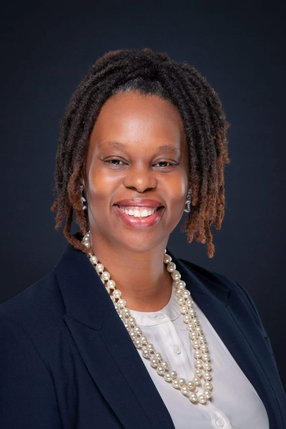 Professional portrait of an African American woman with natural dreadlocks, wearing pearl jewelry, a navy blazer, and white blouse, smiling against a dark background.