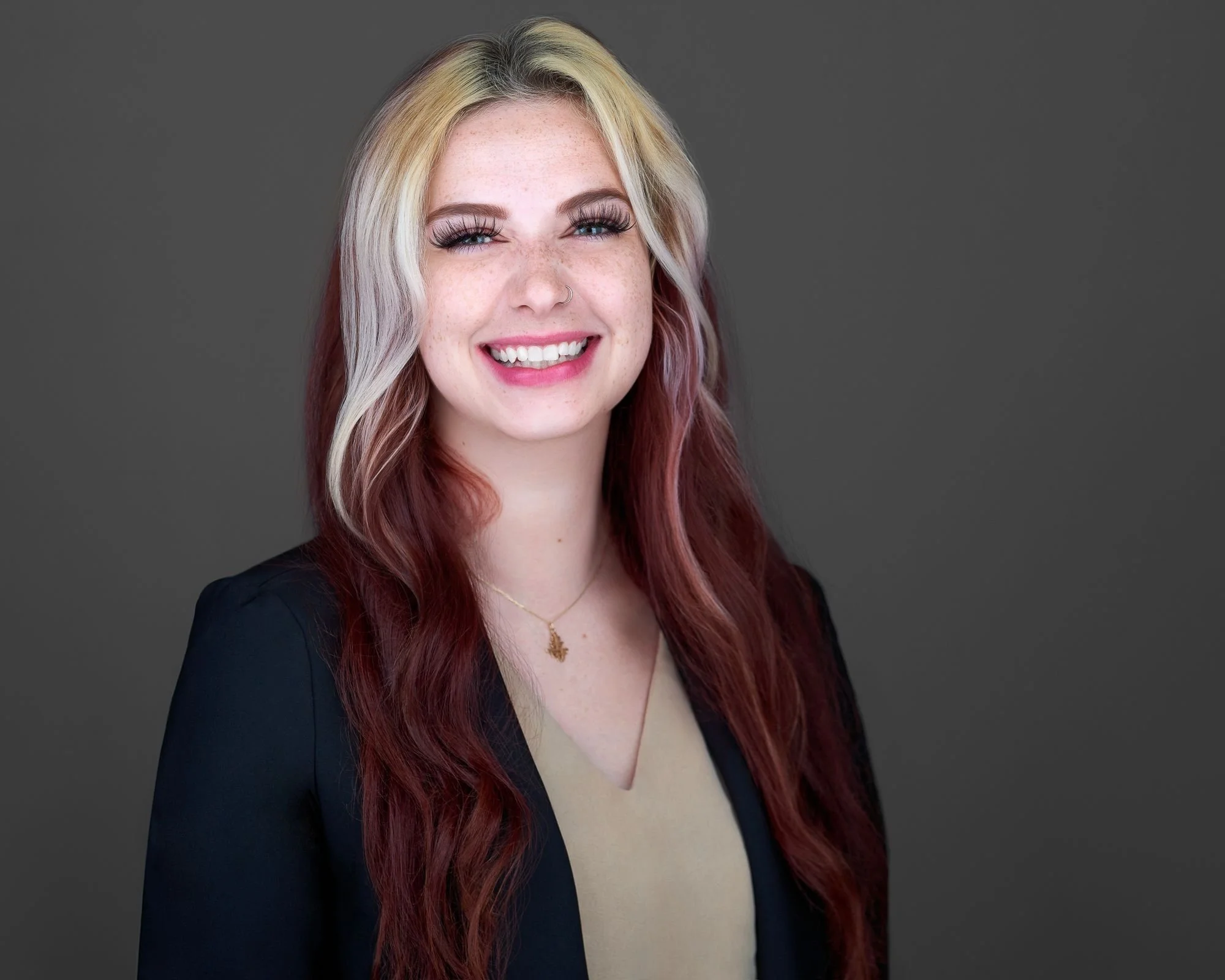 A young woman with long, wavy, multi-colored hair smiling against a dark gray background.