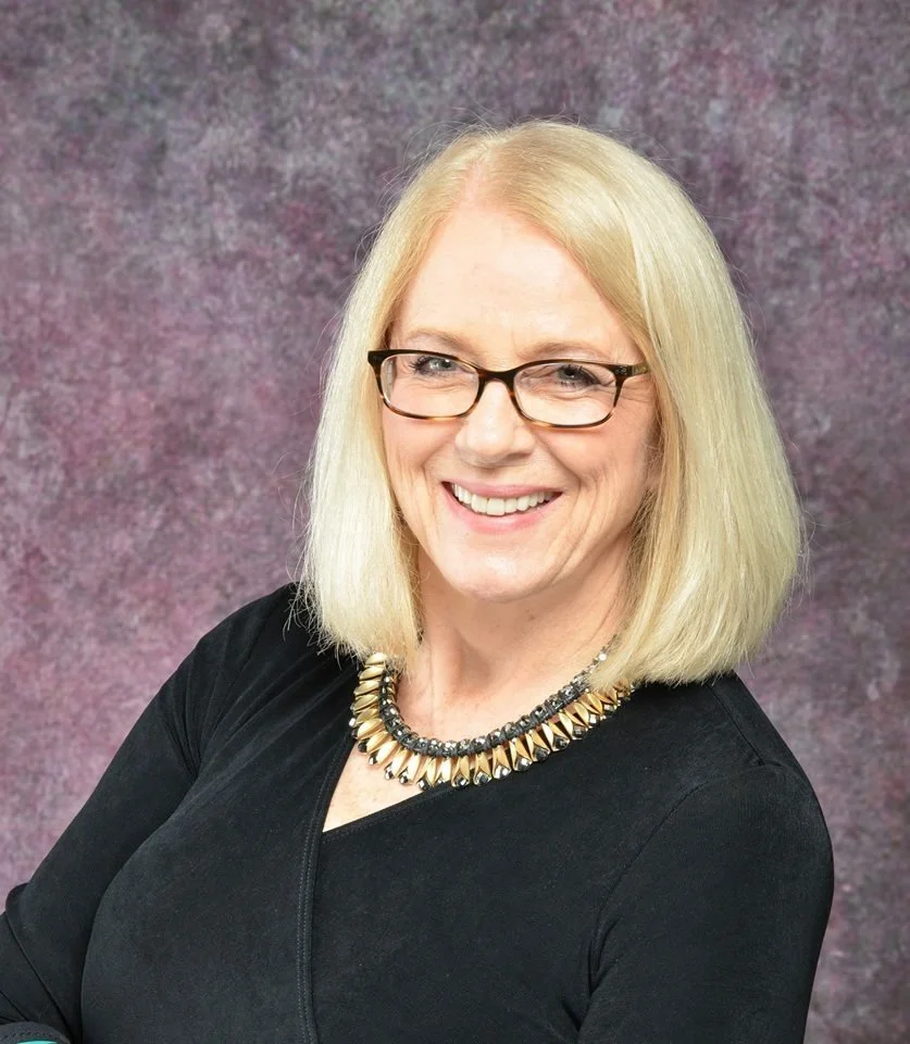 A smiling blonde woman wearing glasses and a black top, with a layered necklace, standing in front of a textured purple and gray background.