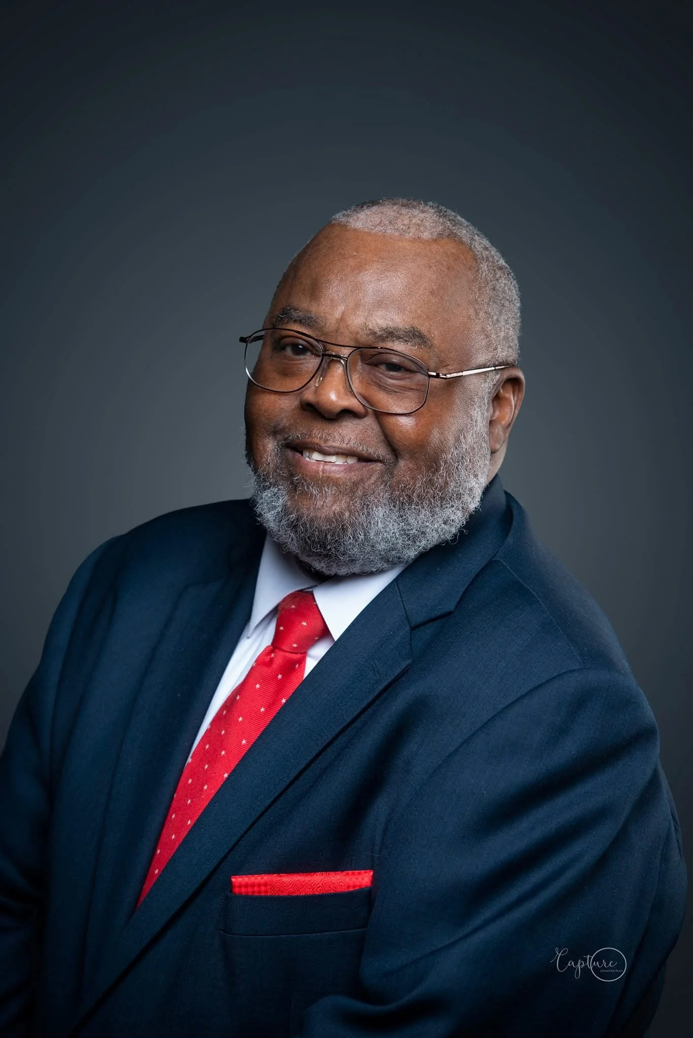 Portrait of a smiling Black man with gray hair and beard wearing glasses, a navy blue suit, white shirt, red tie with white polka dots, and red pocket square, against a dark gray background.