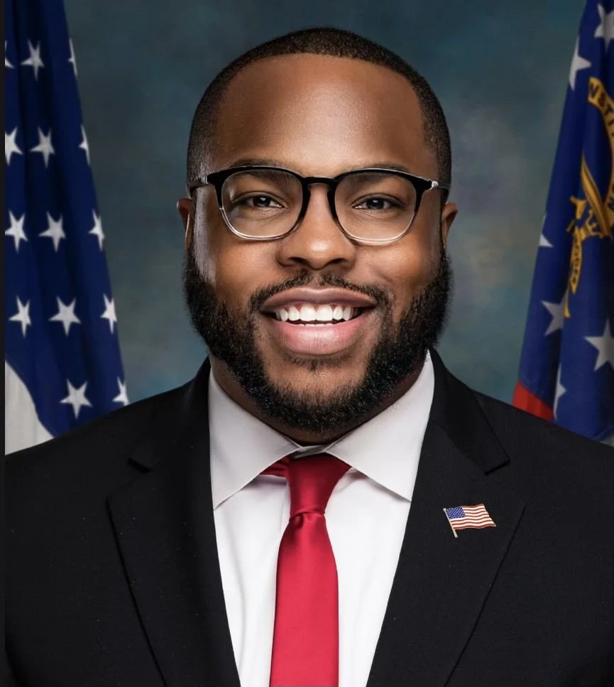 Portrait of a man in a suit with glasses, smiling, with American flags in the background.