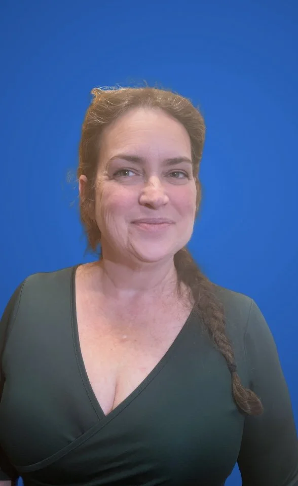 A woman with reddish-brown hair styled in a loose braid, smiling, wearing a dark V-neck top, standing against a solid blue background.