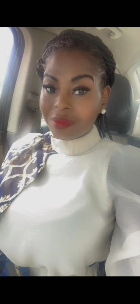 A woman sitting in a car, wearing a white blouse with a decorative navy and gold scarf, pearl earrings, red lipstick, and styled hair.