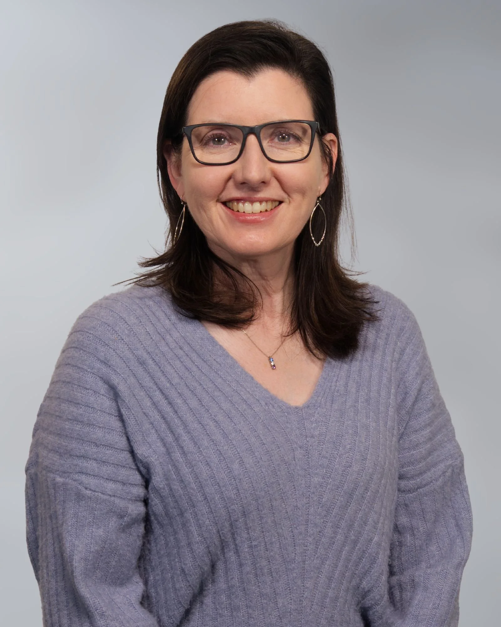A woman with shoulder-length curly brown hair, wearing glasses, smiling, dressed in a dark green V-neck sweater, standing against a light gradient background.