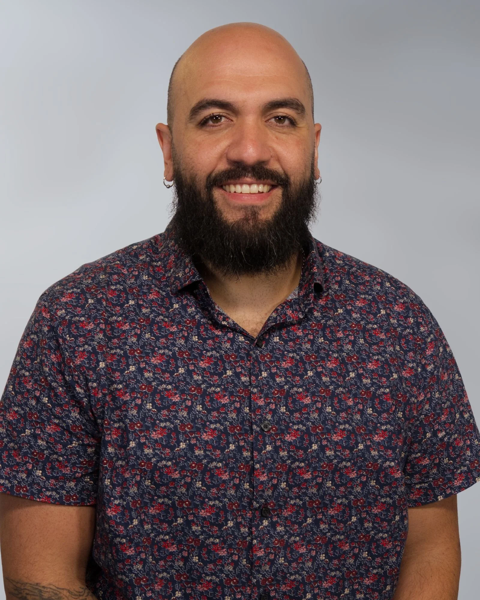 A smiling man with a bald head, full beard, wearing a dark blue button-up shirt with small white dots, standing against a plain beige background.