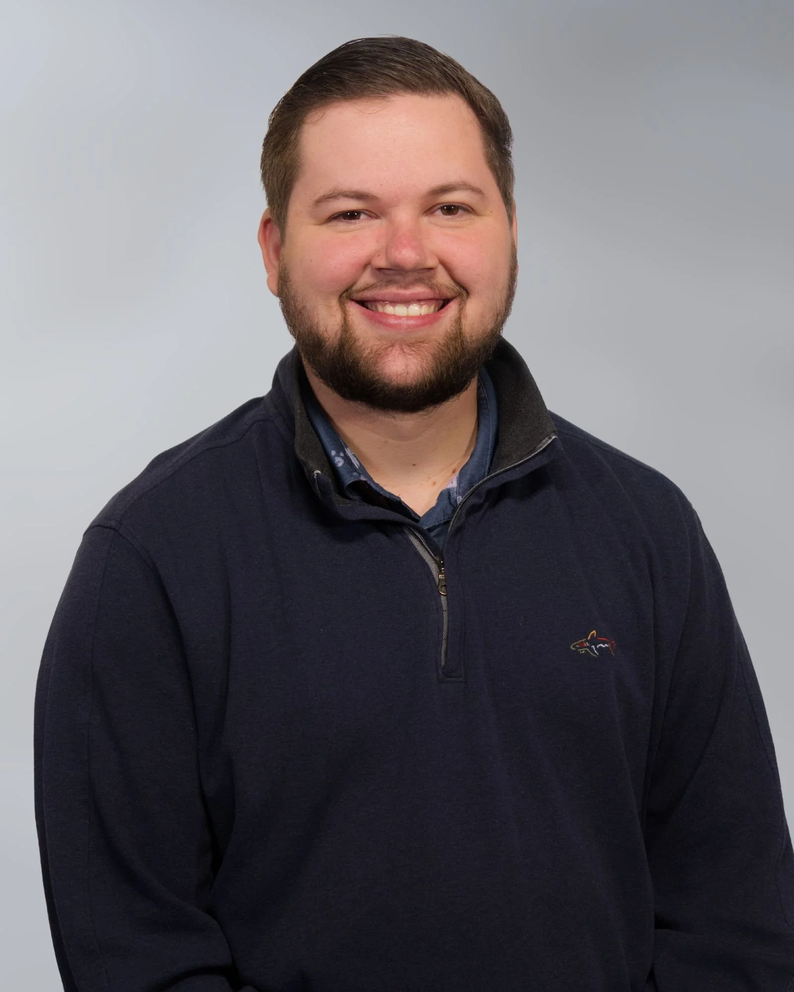 Man with a beard smiling, wearing a casual jacket and a t-shirt with visible text, grey background.