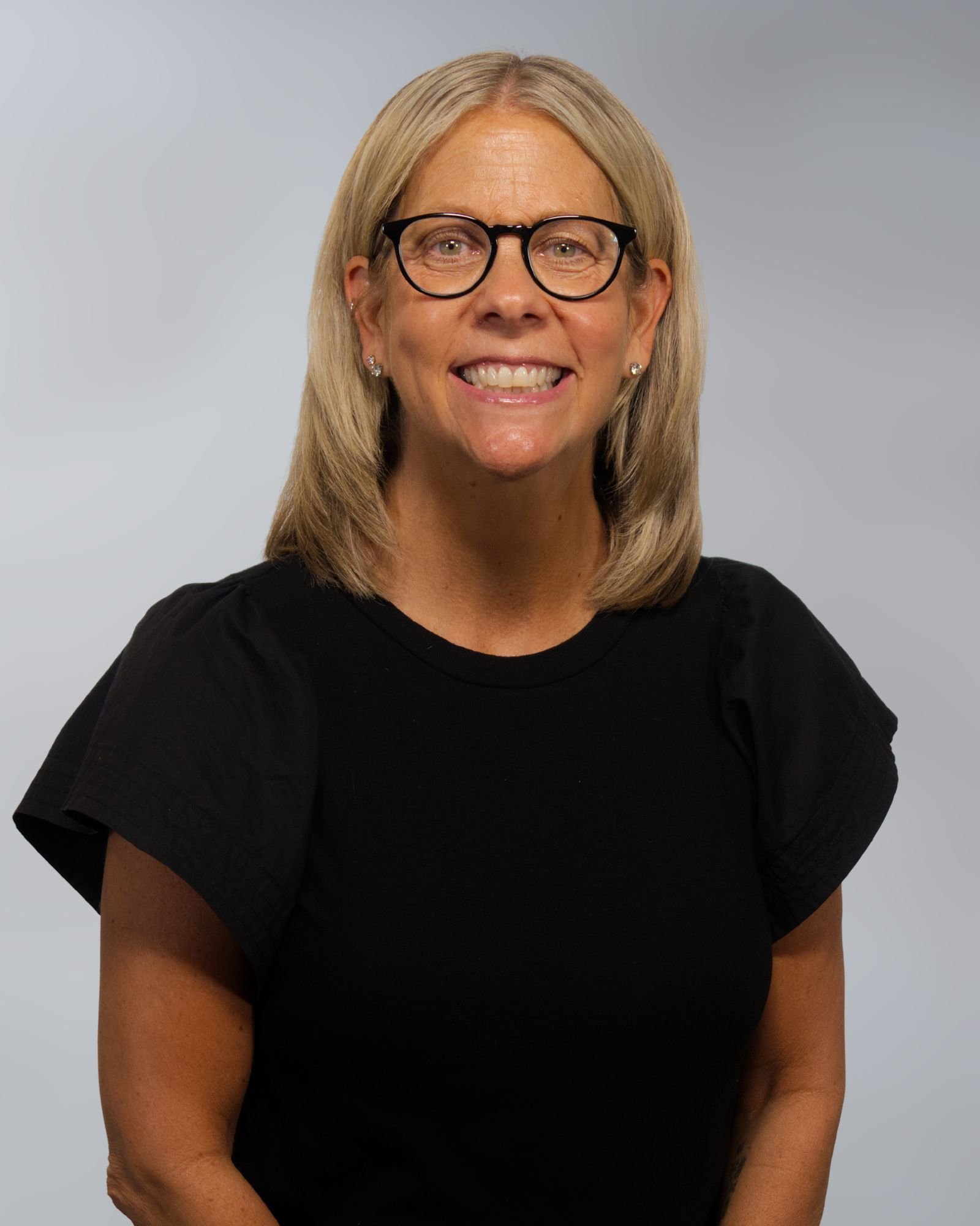 A woman with blonde hair, glasses, and a maroon top, smiling at the camera with a plain light background.