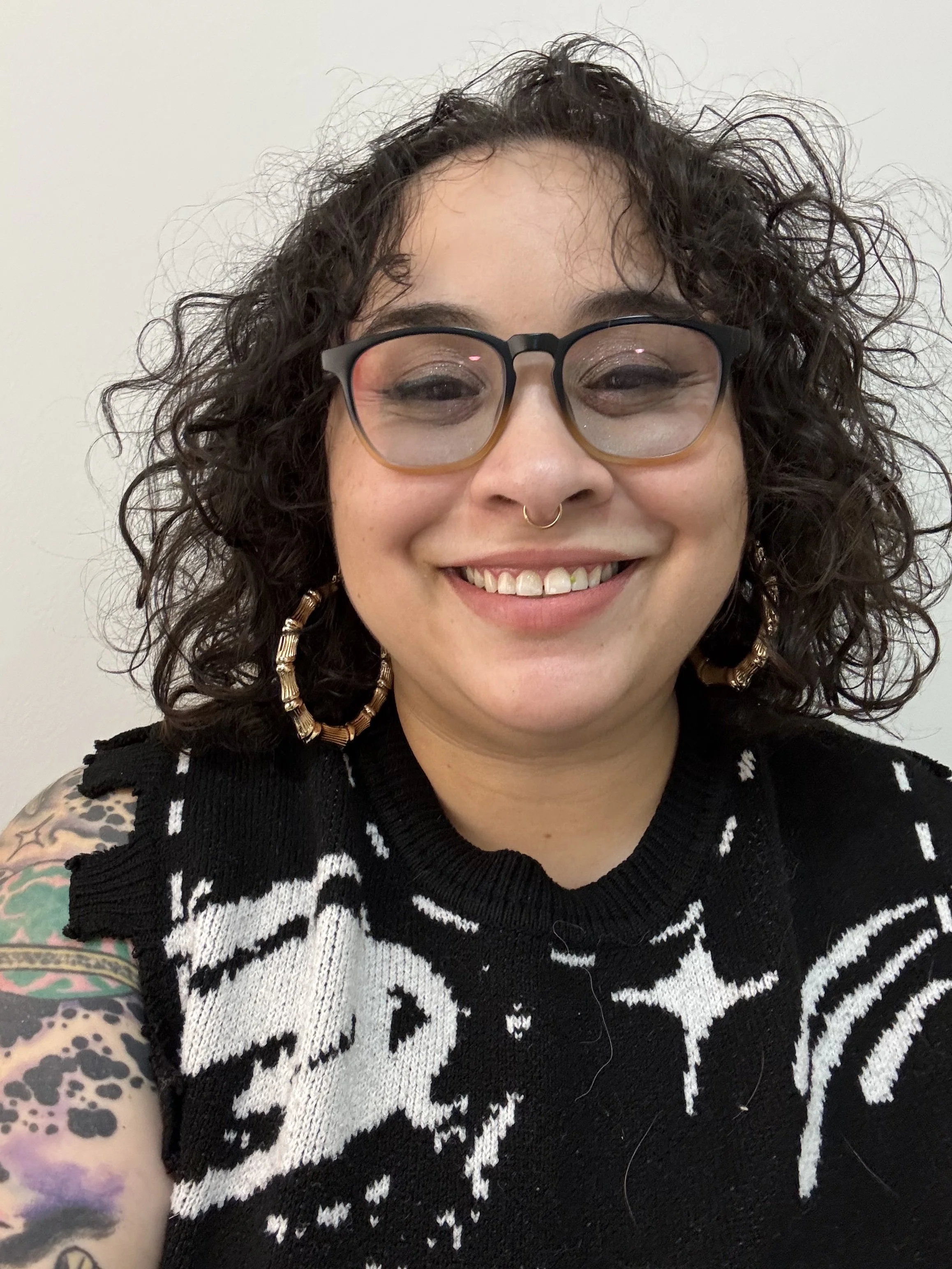 Close-up photo of a woman with dark curly hair, wearing black glasses, gold hoop earrings, a nose ring, and a black and white patterned sweater, smiling and showing her teeth, with a white background.