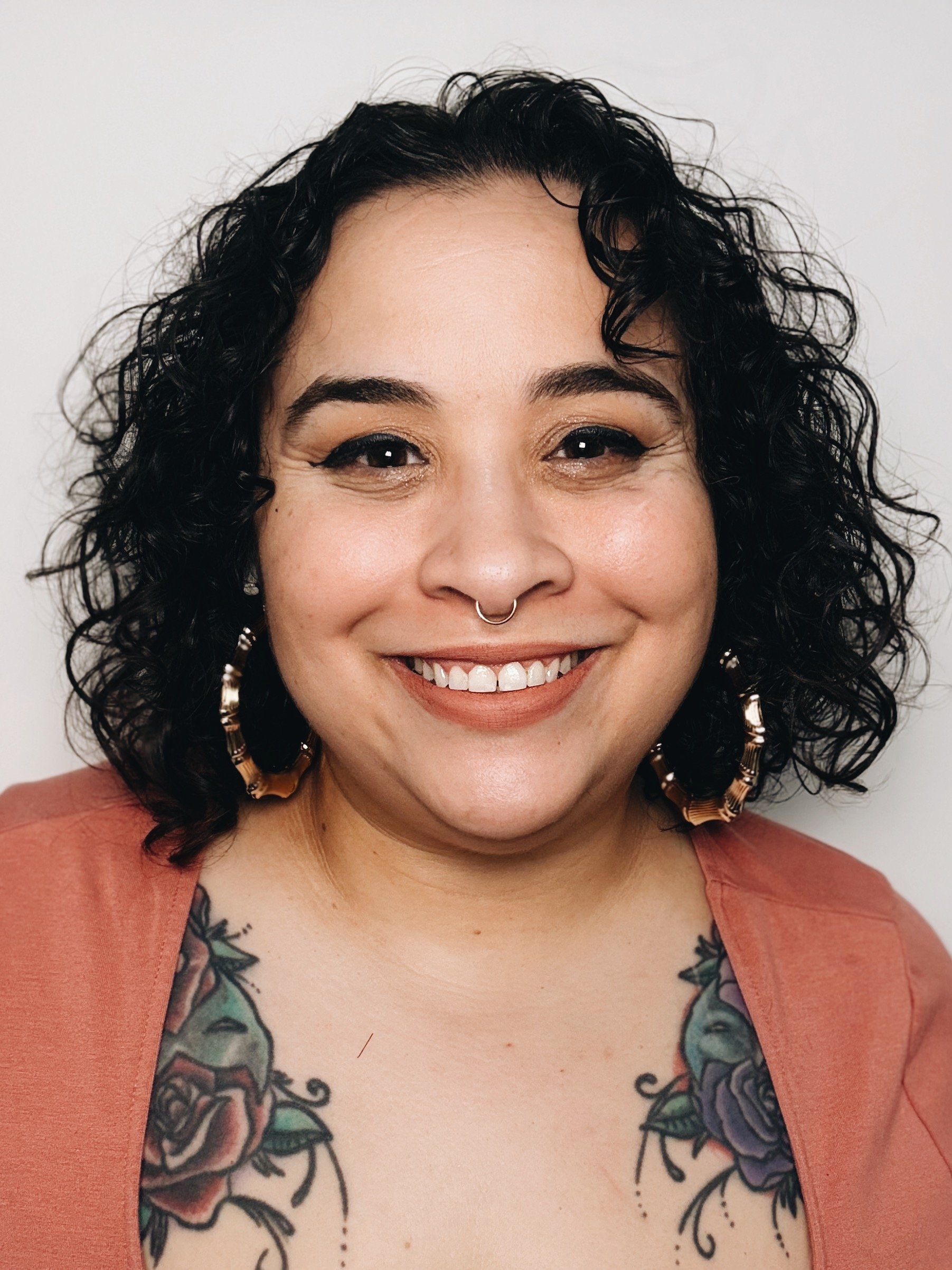 Close-up of a smiling woman with curly black hair, wearing a septum piercing, large hoop earrings, and a tattoo of roses on her chest. She is dressed in a rust-colored top.