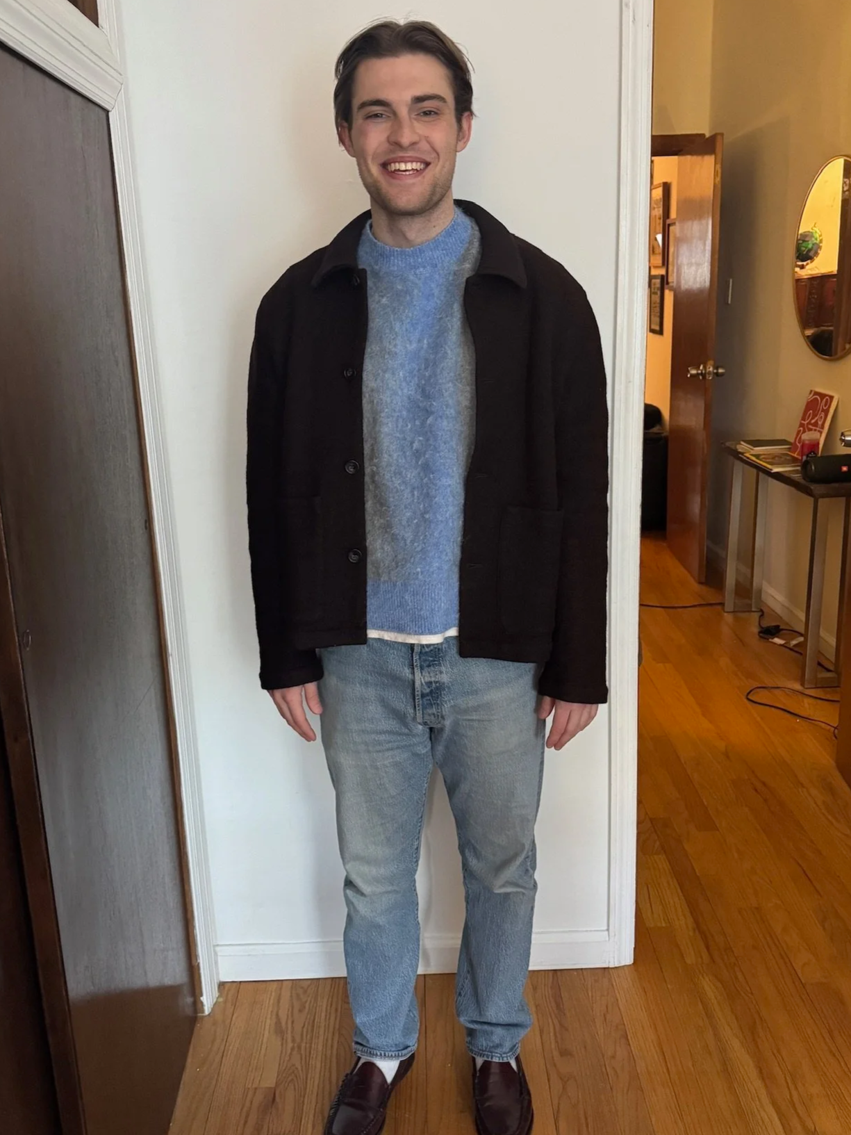 A young man standing indoors, smiling, wearing a black jacket over a blue sweater, light blue jeans, and dark brown shoes.