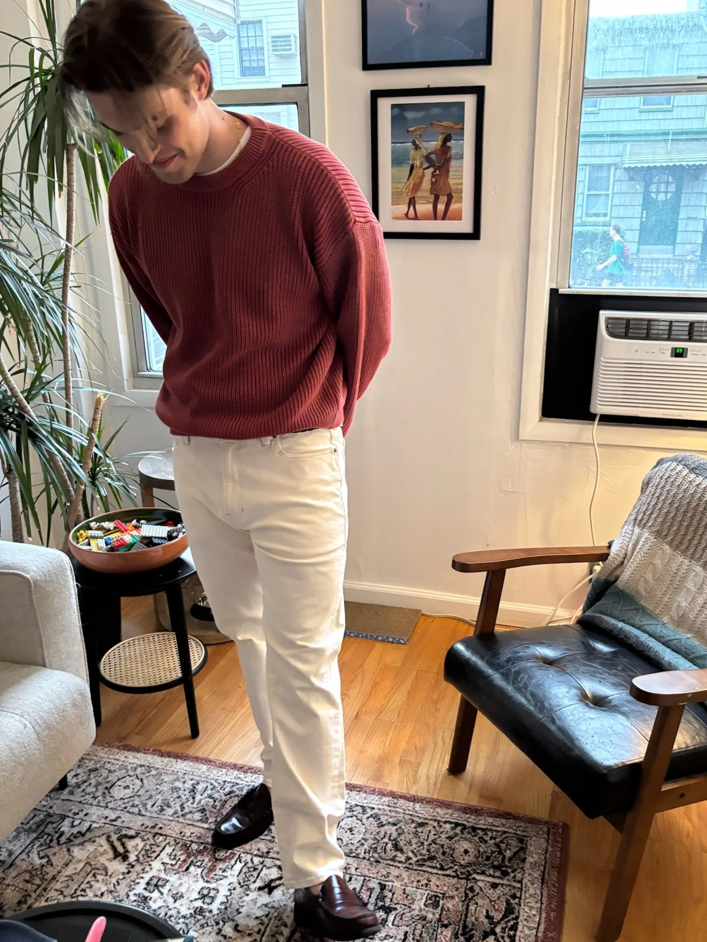 A young man with light skin and brown hair styled in a messy look, wearing a red sweater, white pants, and brown shoes.