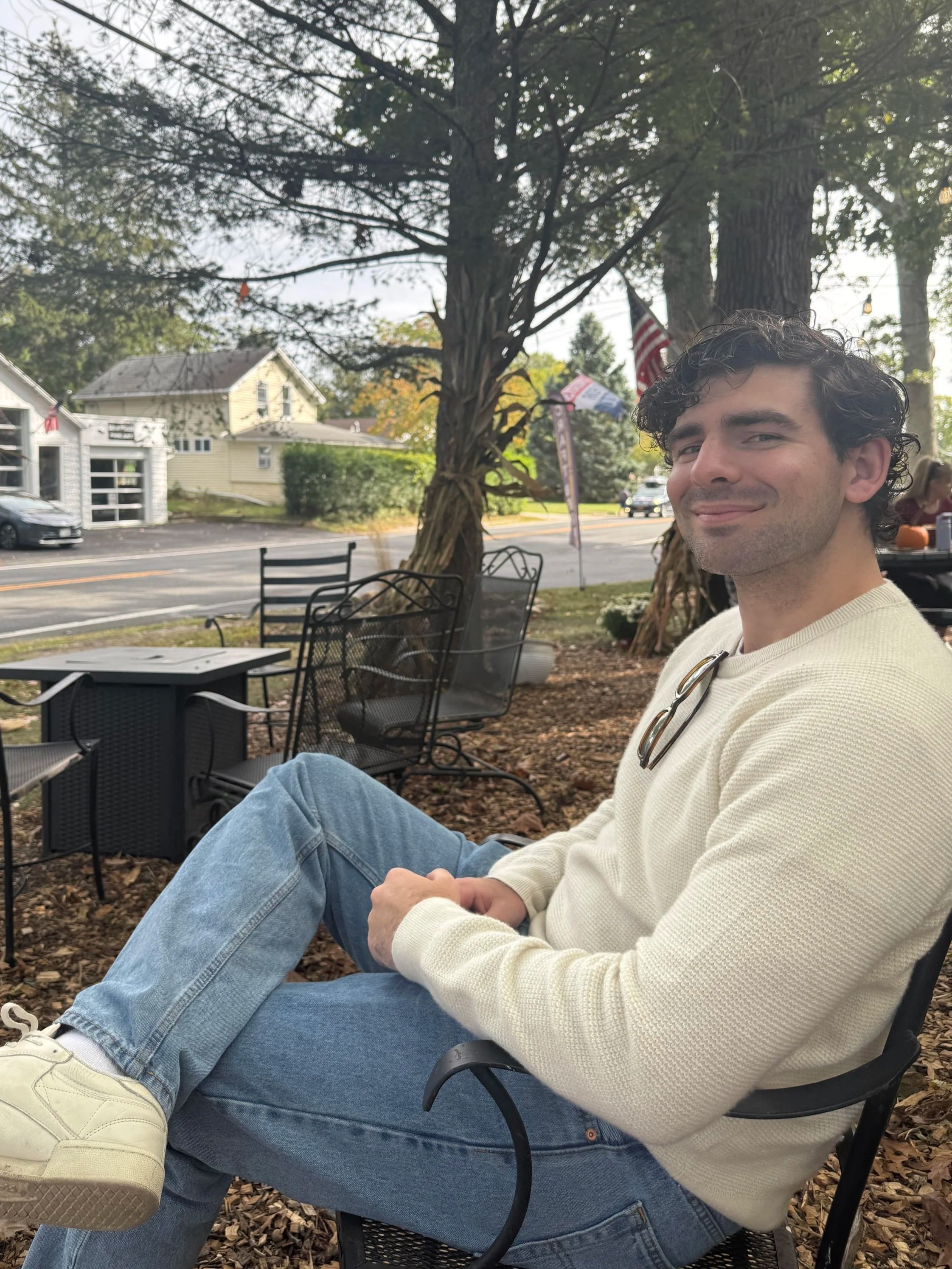 A young man sitting outside in a chair on a patio with trees, a street, and houses in the background, smiling at the camera.