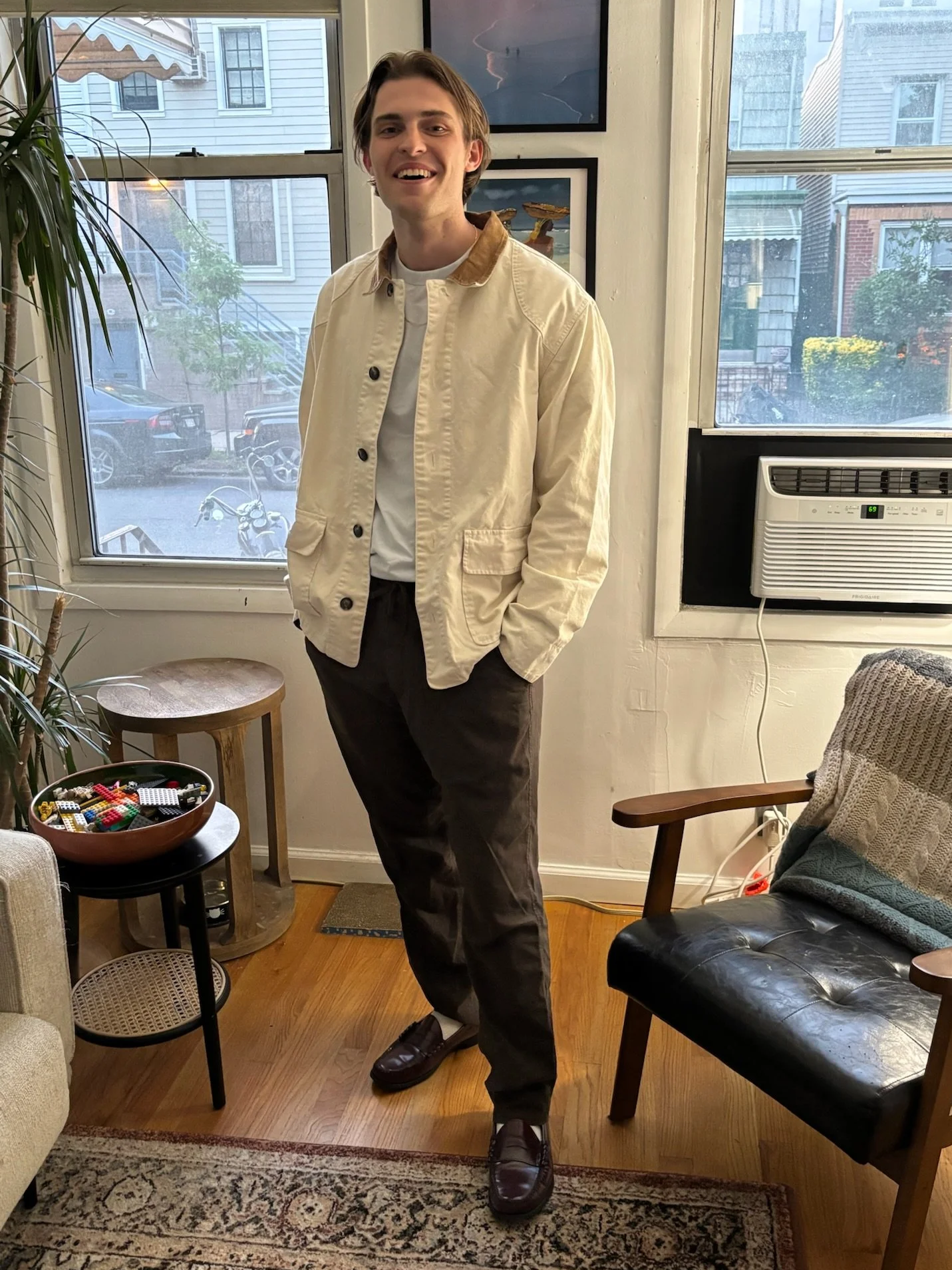 A young man with light brown hair is standing inside a cozy room, smiling at the camera. He is wearing a cream-colored jacket with a brown collar, a white t-shirt, dark pants, and brown loafers.