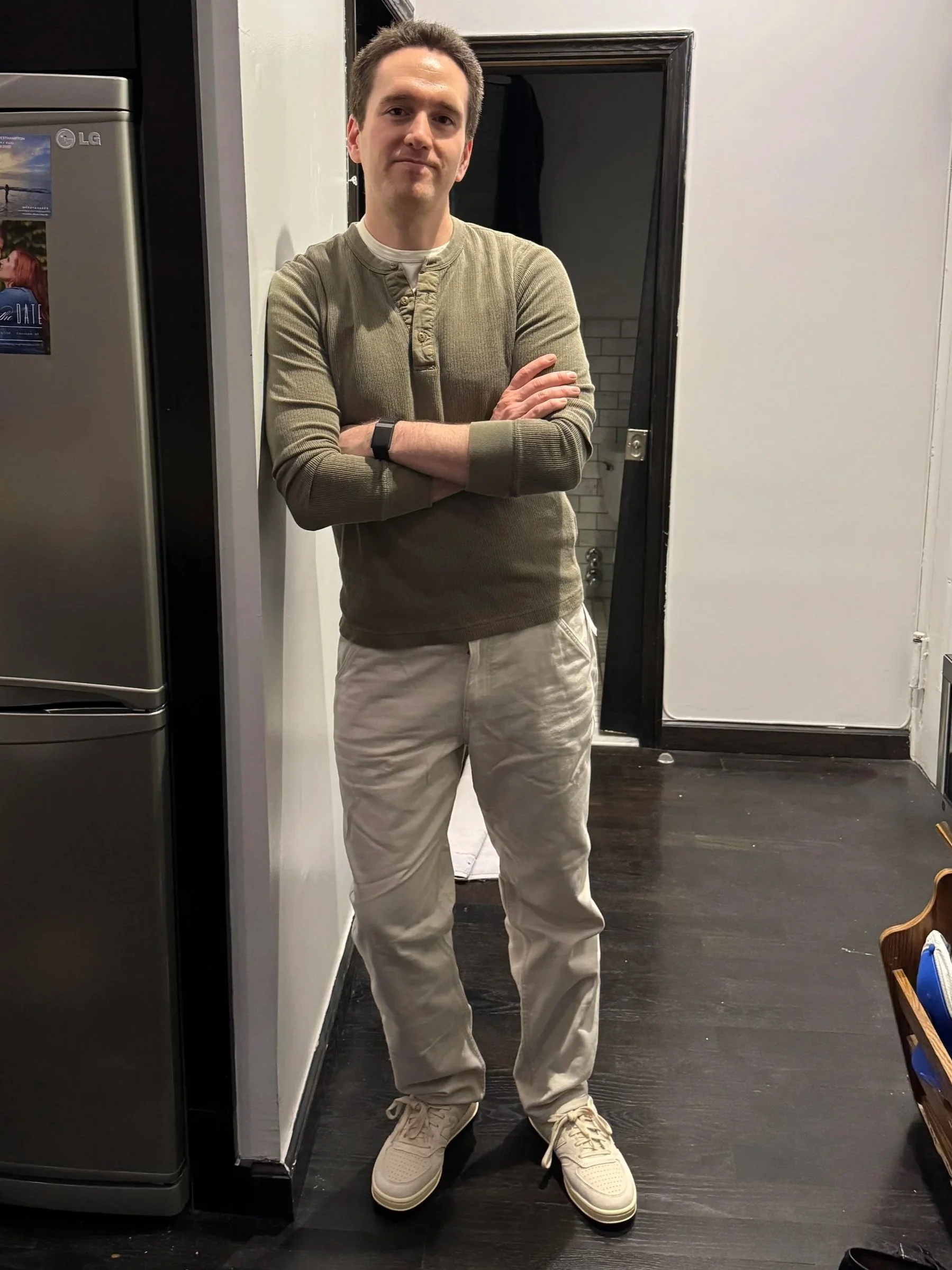 A man standing indoors with arms crossed, wearing a tan sweater, beige pants, and white sneakers, in a kitchen or laundry area.