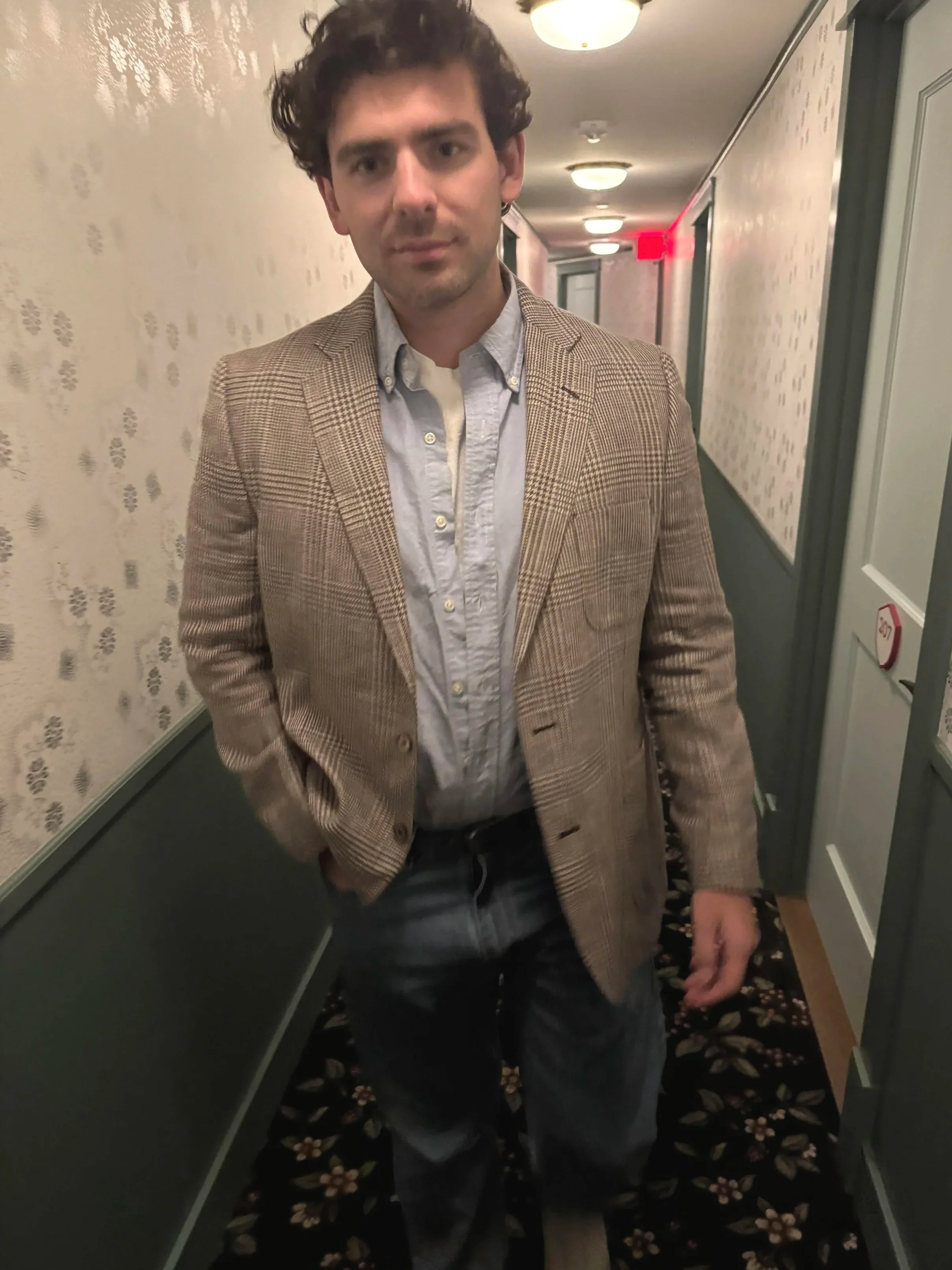 A man with curly dark hair wearing a beige plaid blazer, light blue button-up shirt, and dark jeans is standing in a hallway.