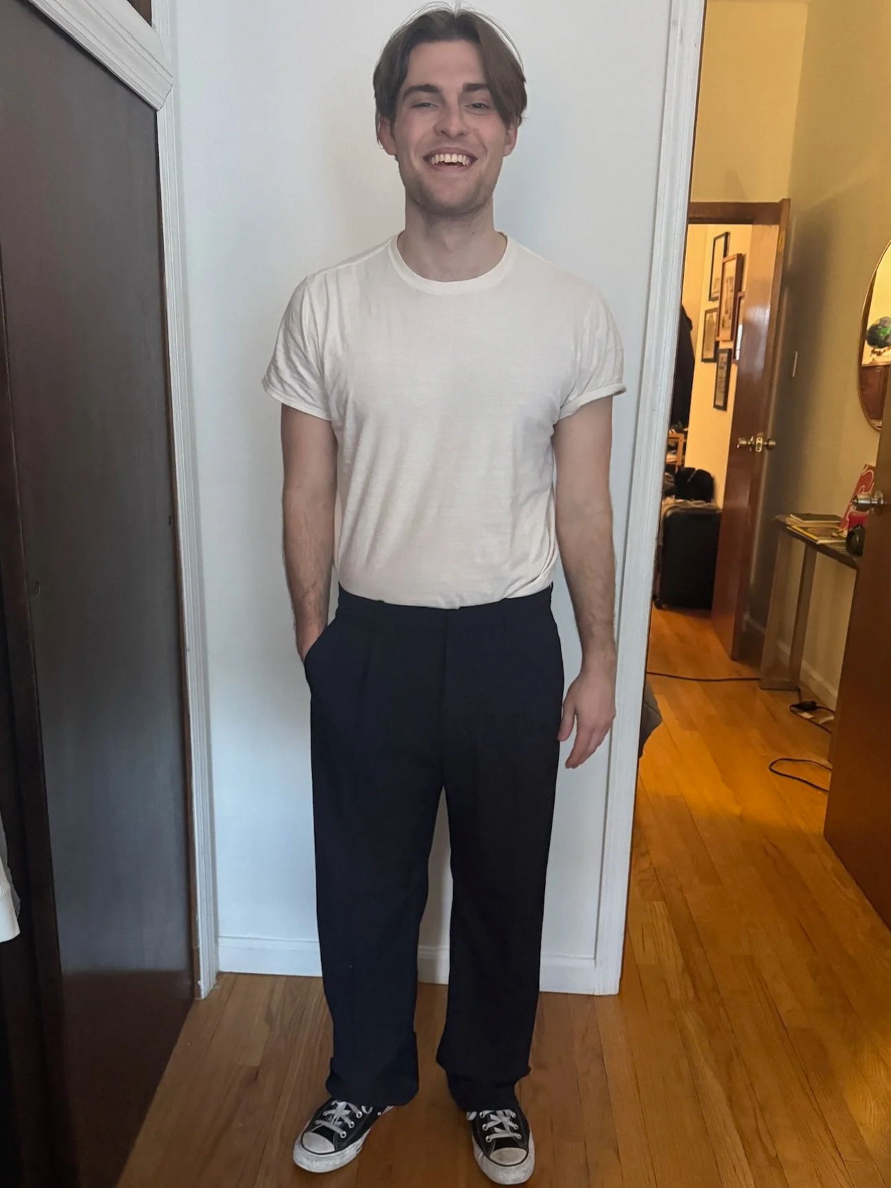 A young man with brown hair, wearing a white t-shirt, black pants, and black sneakers, standing indoors and smiling.