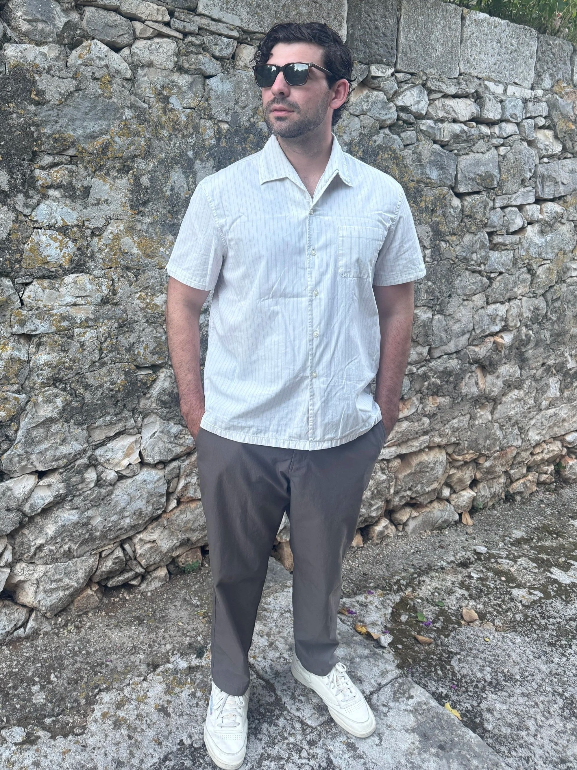A man with dark, wavy hair and sunglasses standing outdoors against a stone wall, wearing a short-sleeved, light-colored button-up shirt, dark pants, and white sneakers.