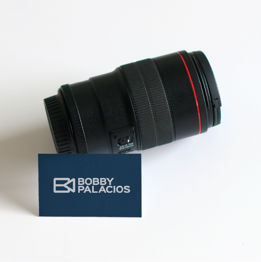A camera lens with a black and red design is positioned next to a blue business card featuring the name "Bobby Palacios" and a logo.