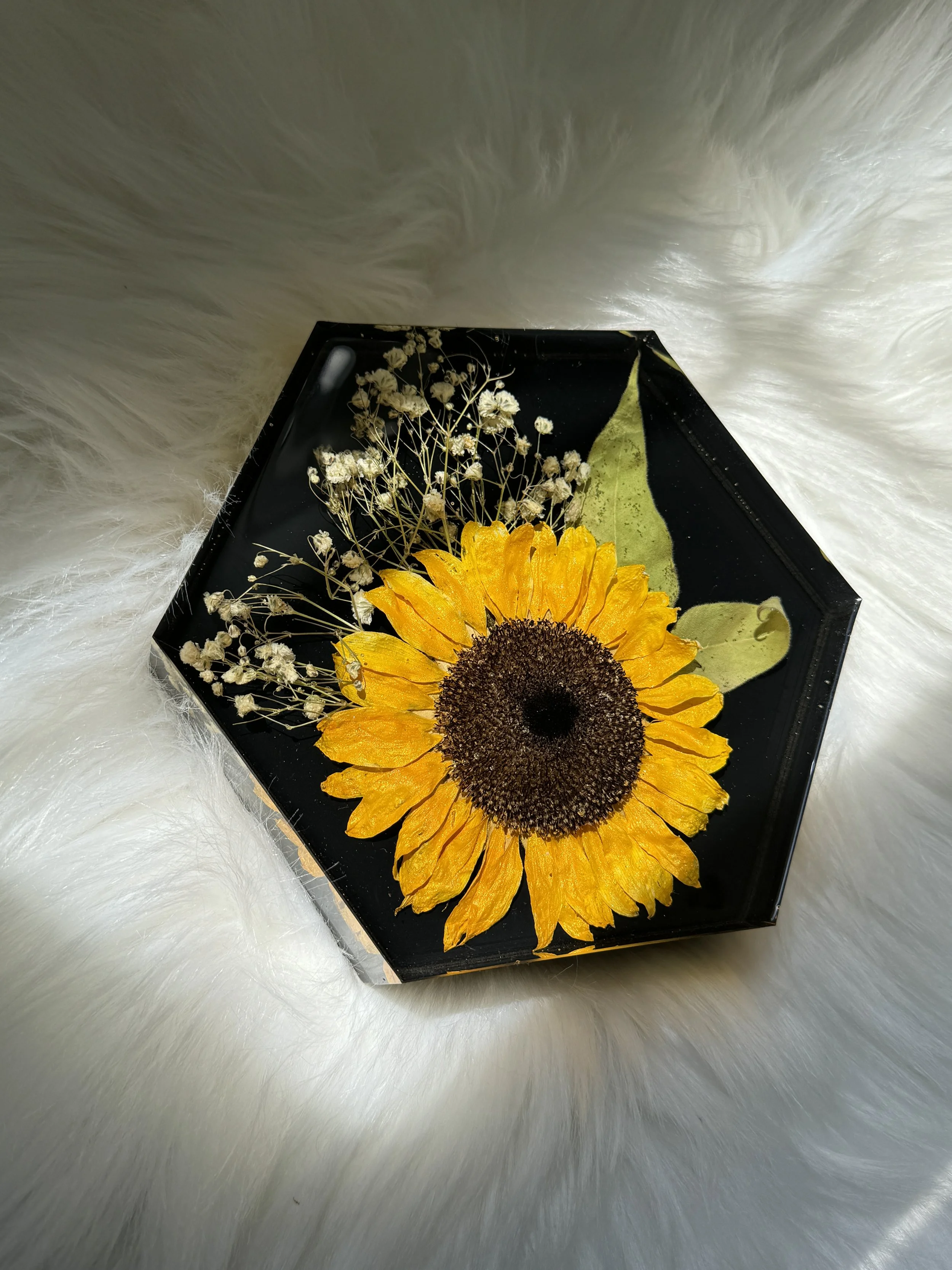 Hexagonal tray with a pressed sunflower, small white flowers, and green leaves on a white fur background.