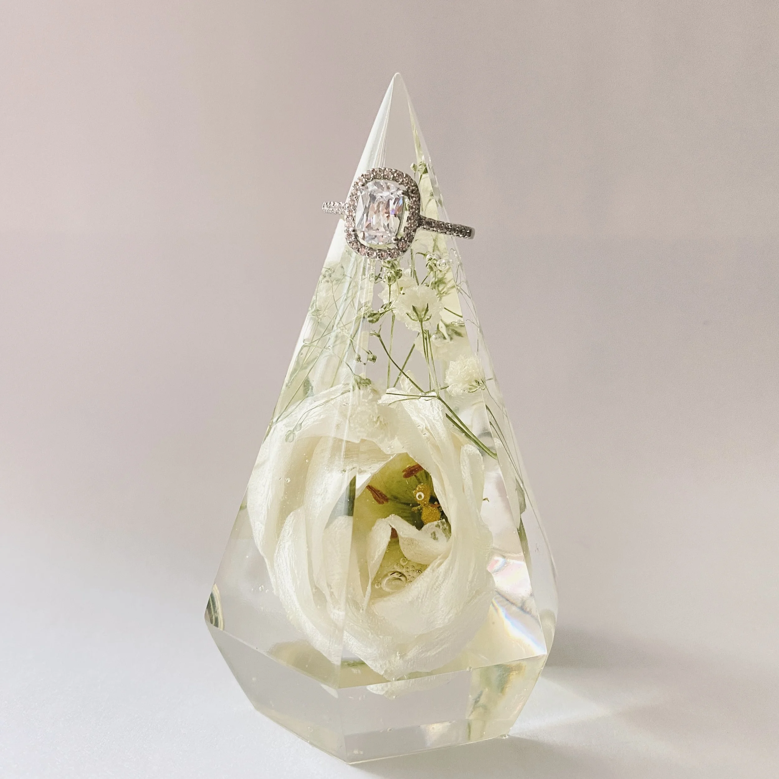 A clear resin pyramid encasing a white rose and small flowers with a diamond halo engagement ring displayed on top against a neutral background.