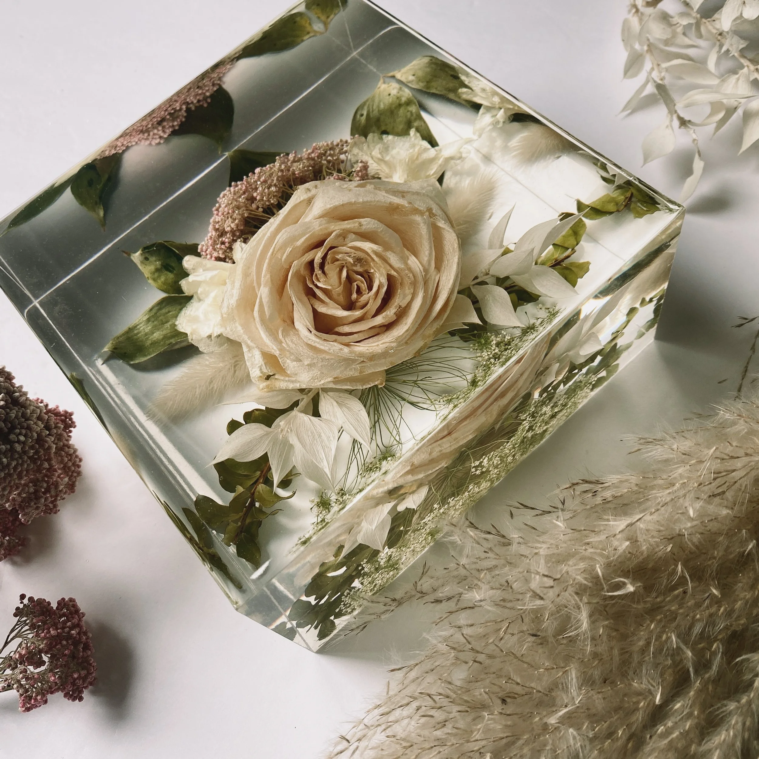 A preserved flower arrangement encased in a clear resin block, featuring a central beige rose with green and white foliage, displayed on a white background. Nearby, there are dried flowers and ornamental grasses.