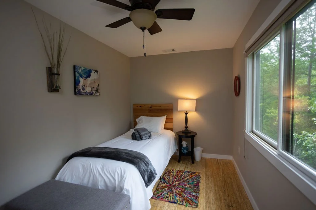 A cozy bedroom with white walls, wooden framing around two windows, and a glass door leading outside, decorated with curtains and a small bed with towels folded on top.