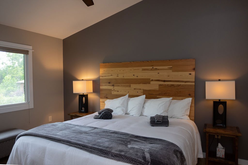 Room with three beds, wooden walls, window, ceiling fan, two lamps, and framed pictures.