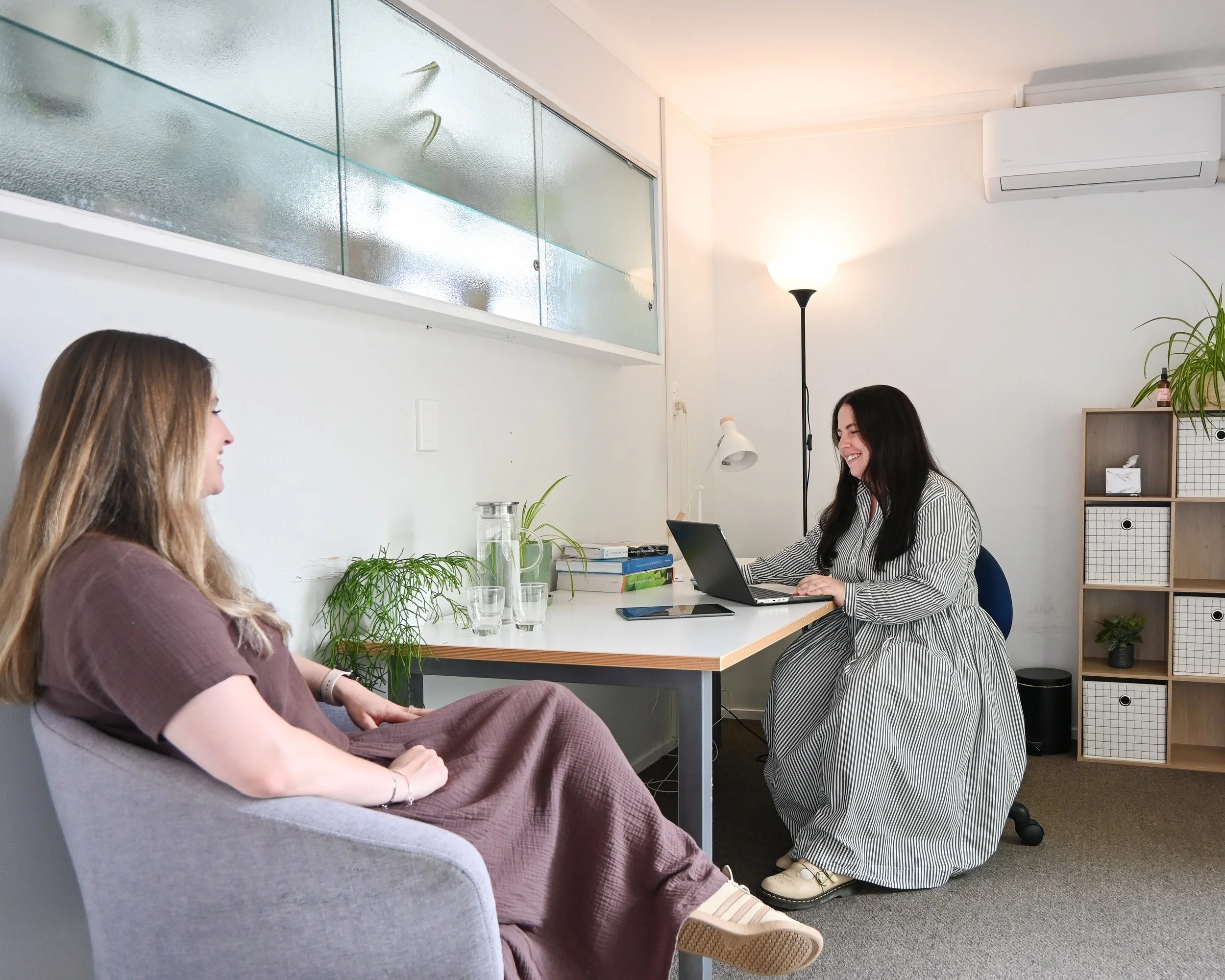 A woman sitting on a gray armchair talking to a woman working on a laptop at a white desk in an office, with a window and green plants in the background.