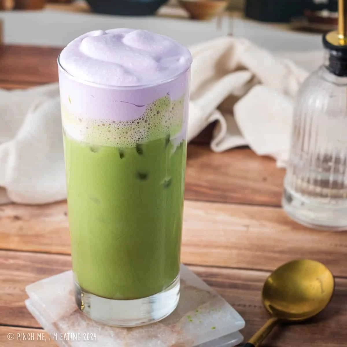 A tall glass filled with a bright green matcha latte topped with purple foam, placed on a wooden table with a gold spoon nearby.