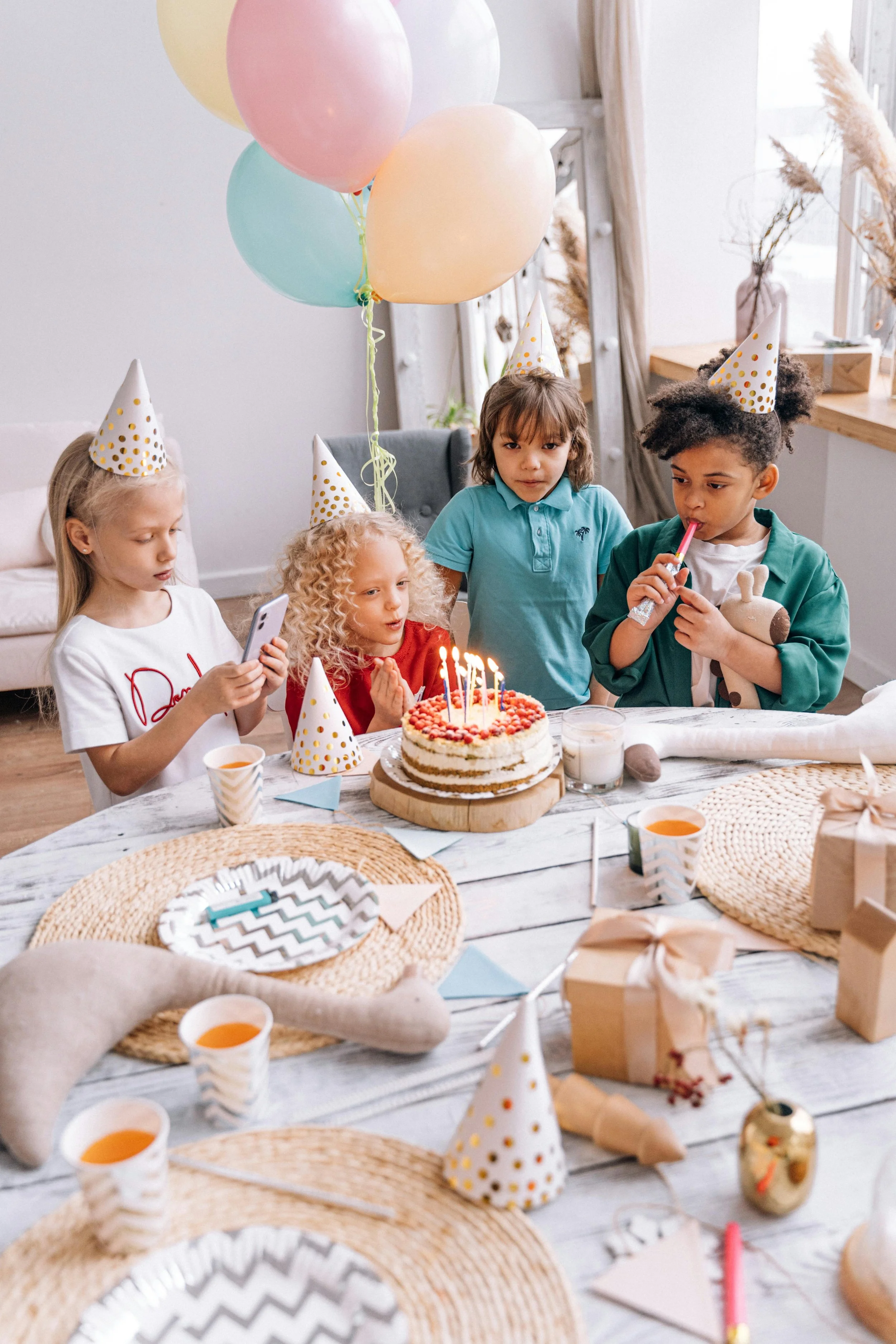 Children celebrating a birthday with a cake, party hats, balloons, and gifts at a decorated table.