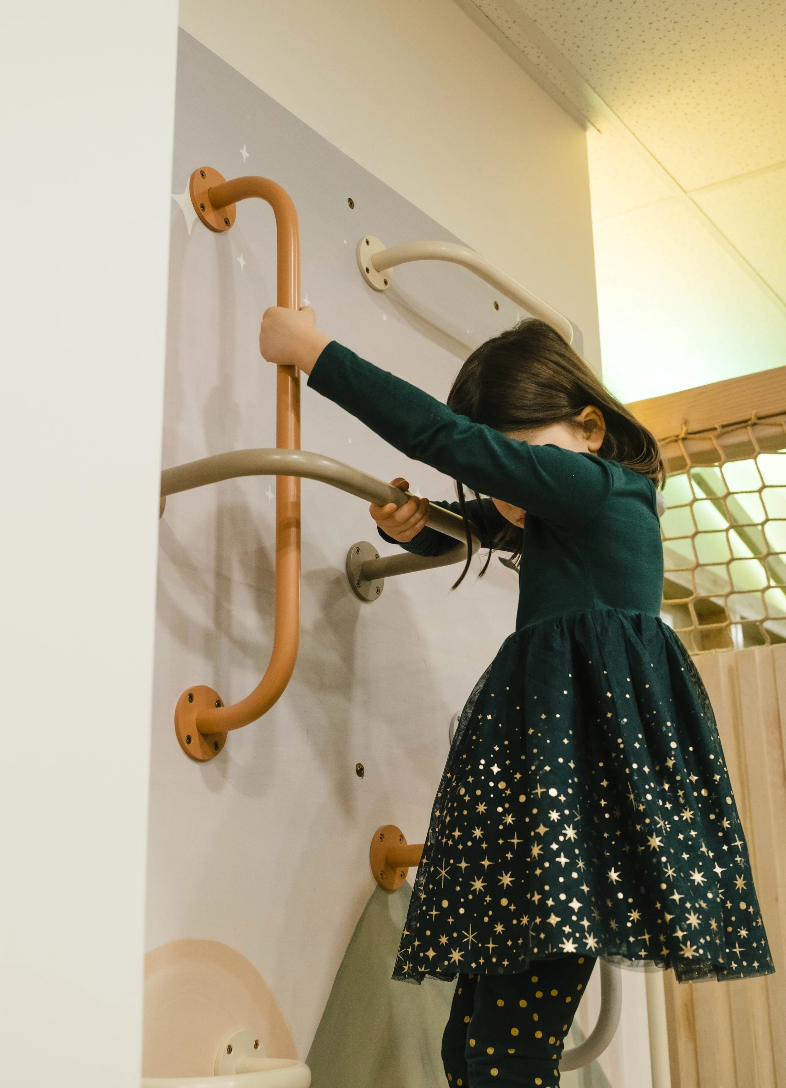 A young girl is climbing a wall-mounted play structure with colorful bars in an indoor play area.