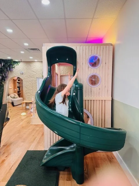 A young girl playing on a green indoor spiral slide with a wooden play structure behind her in a play area.