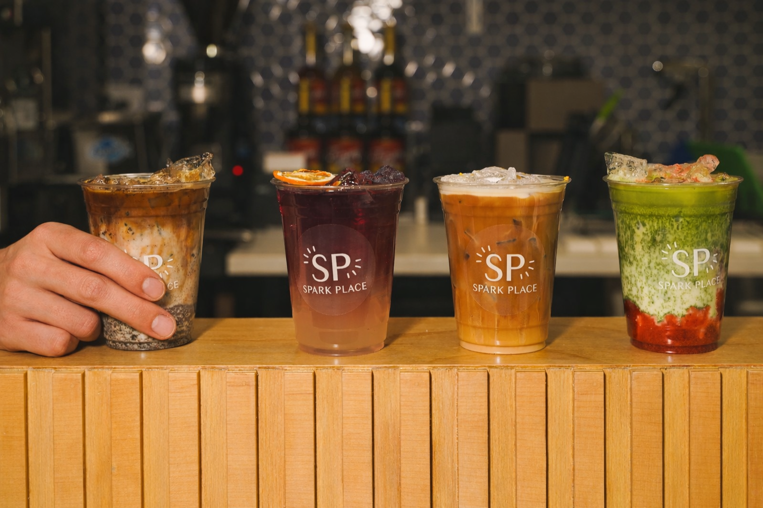 Four colorful iced drinks in clear plastic cups with 'Spark Place' logo on a wooden counter, in a barista or cafe setting.