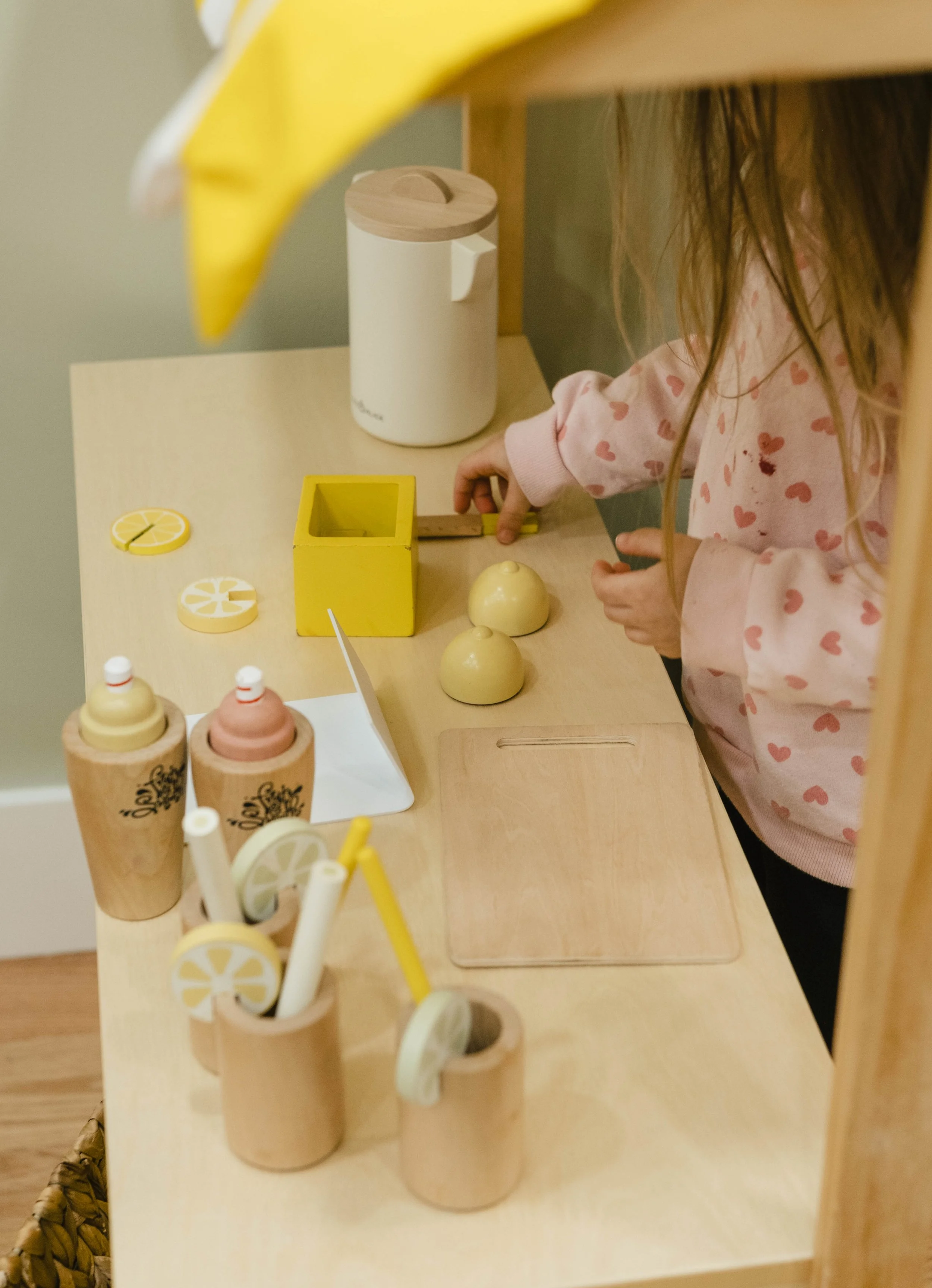 A child in a pink sweatshirt with red hearts playing with wooden and plastic lemon-shaped toys on a light-colored table, with cups that look like ice cream cones and a yellow tissue box, under a yellow lamp.