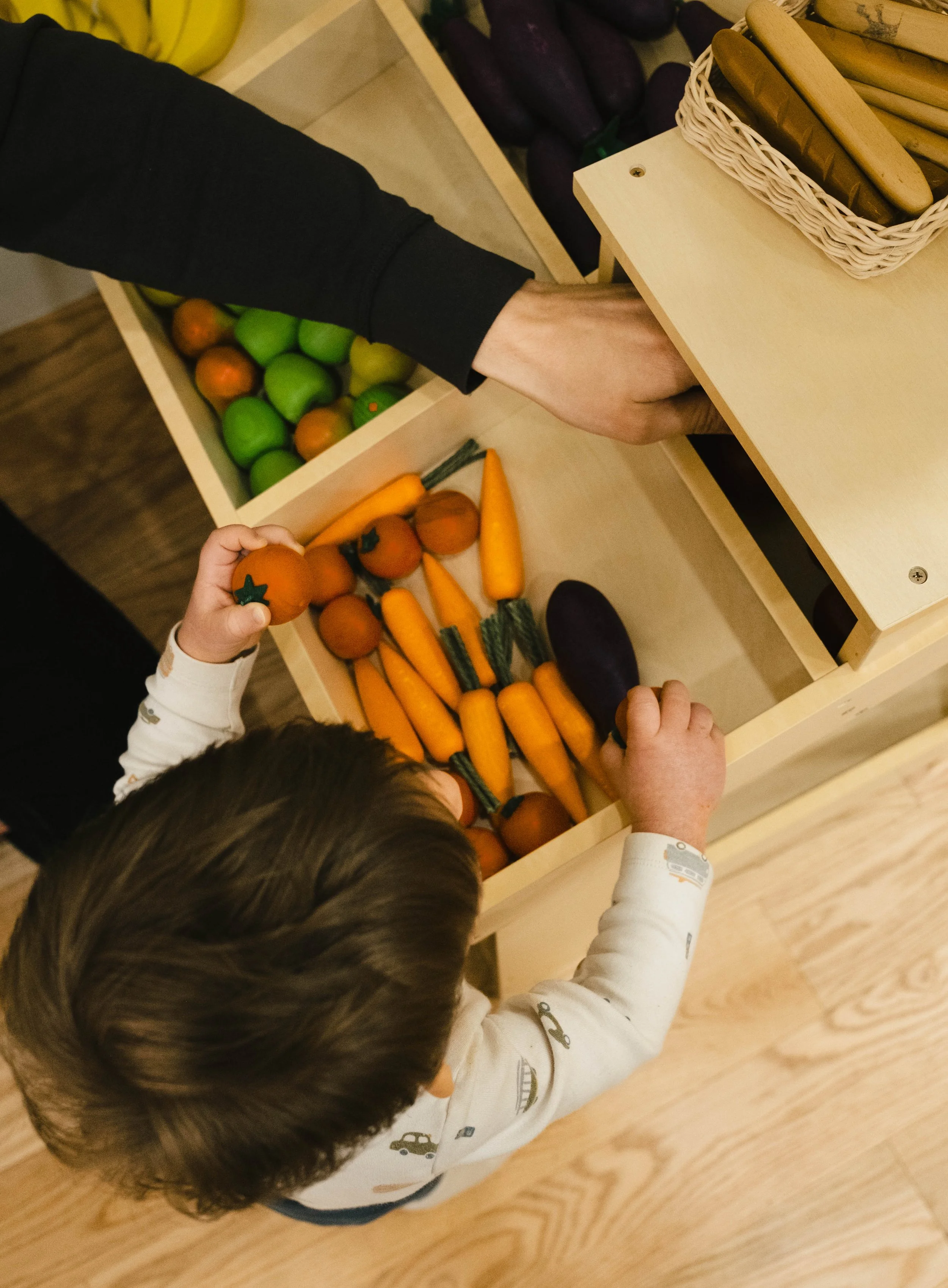A child and an adult are reaching into open vegetable drawers, selecting pretend vegetables including carrots, eggplants, and tomatoes, in a grocery store or play area.