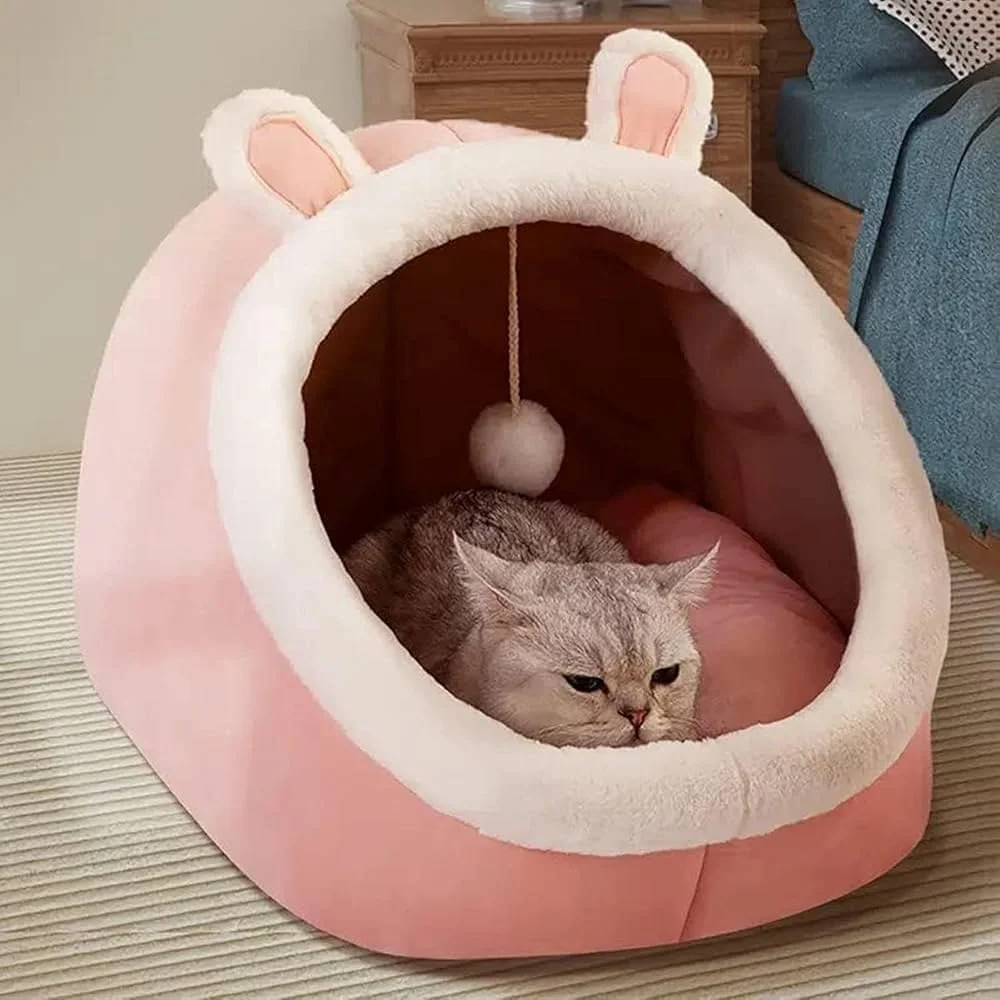 A cat lying inside a pink, plush, bear-shaped pet bed with bear ears and a hanging pom-pom toy.