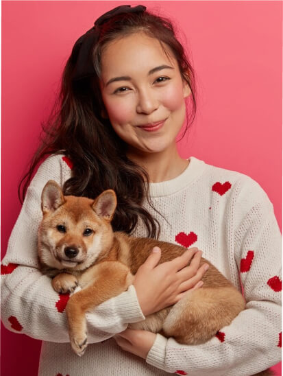 A young woman with long dark hair, wearing a white sweater with red hearts, holding a small brown puppy against a pink background.
