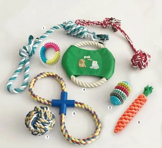 Collection of colorful dog toys including ropes, a rubber ring, a green fabric band with the word 'pet' and cartoon images, a blue rubber ring, a multicolored ball, a fleecy knot, a braided rope with a blue cross, a crocheted carrot, and a textured ball.
