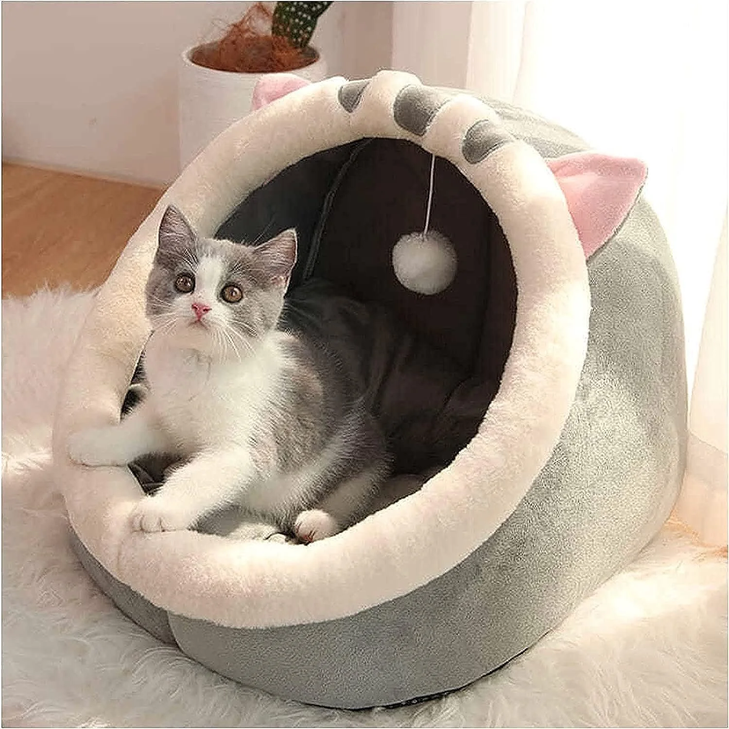 Gray and white kitten sitting inside a cozy cat bed shaped like a cat's head with pink ears and a felt nose, placed on a white fluffy rug.