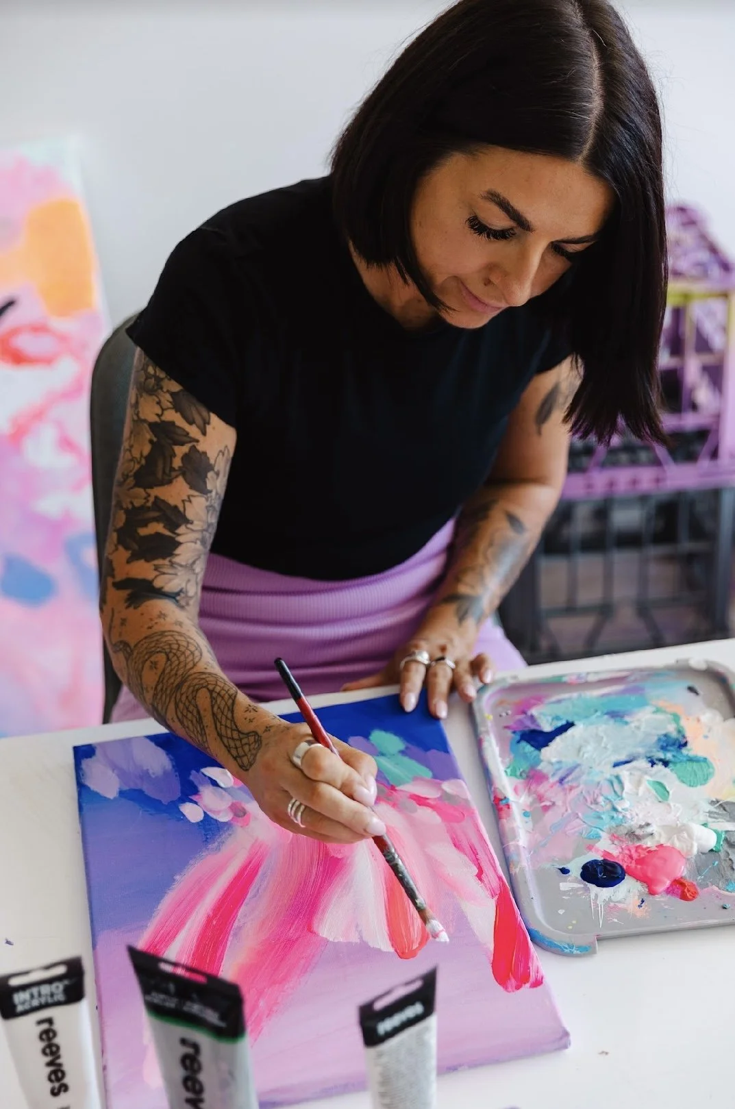 A woman with tattoos on her arms is painting on a canvas with pink, purple, and blue colors, using a paintbrush, in an art studio.