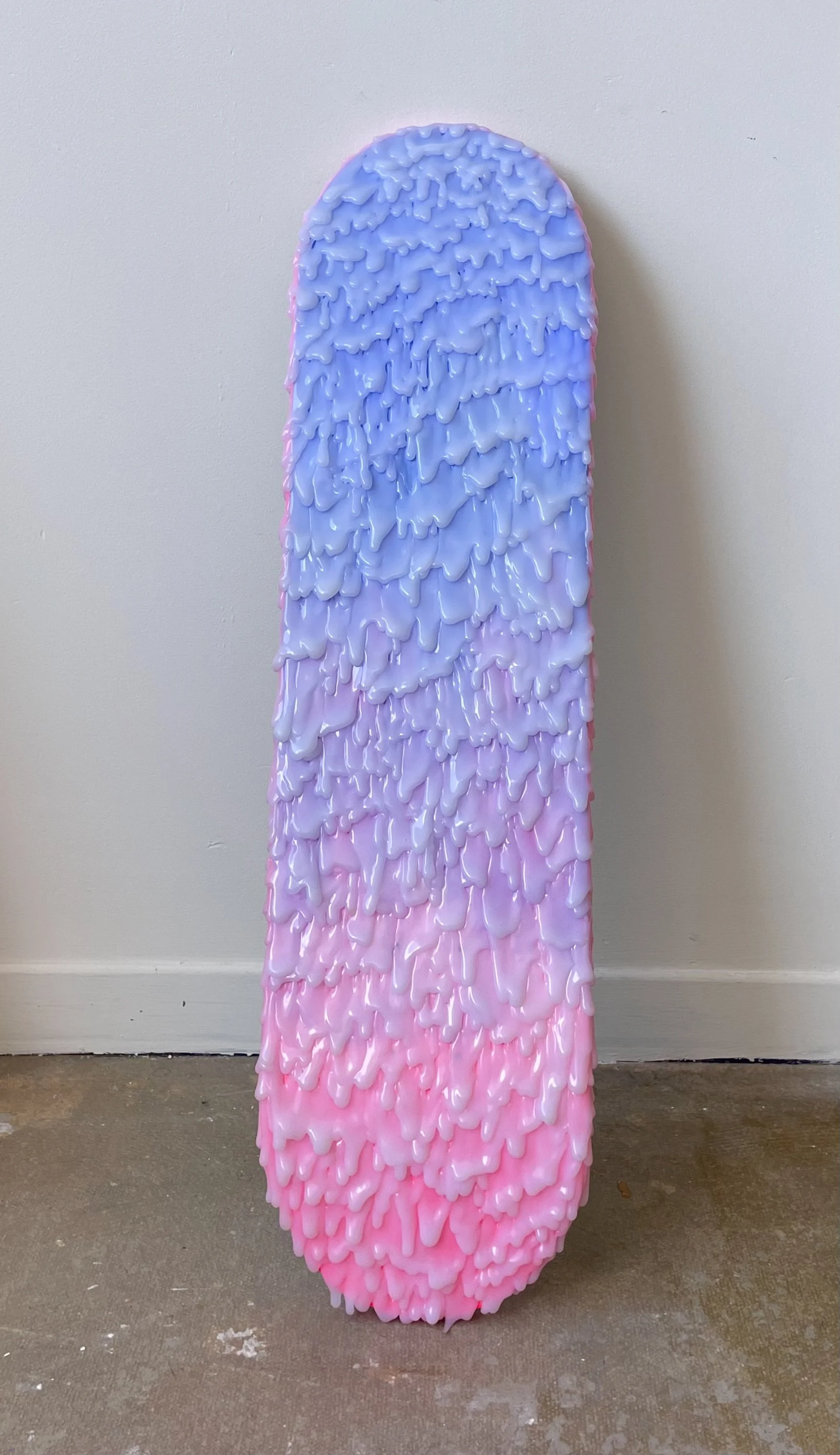 A sculpture resembling a skateboard decorated with textured, dripping paint in shades of blue, purple, and pink standing against a plain wall.