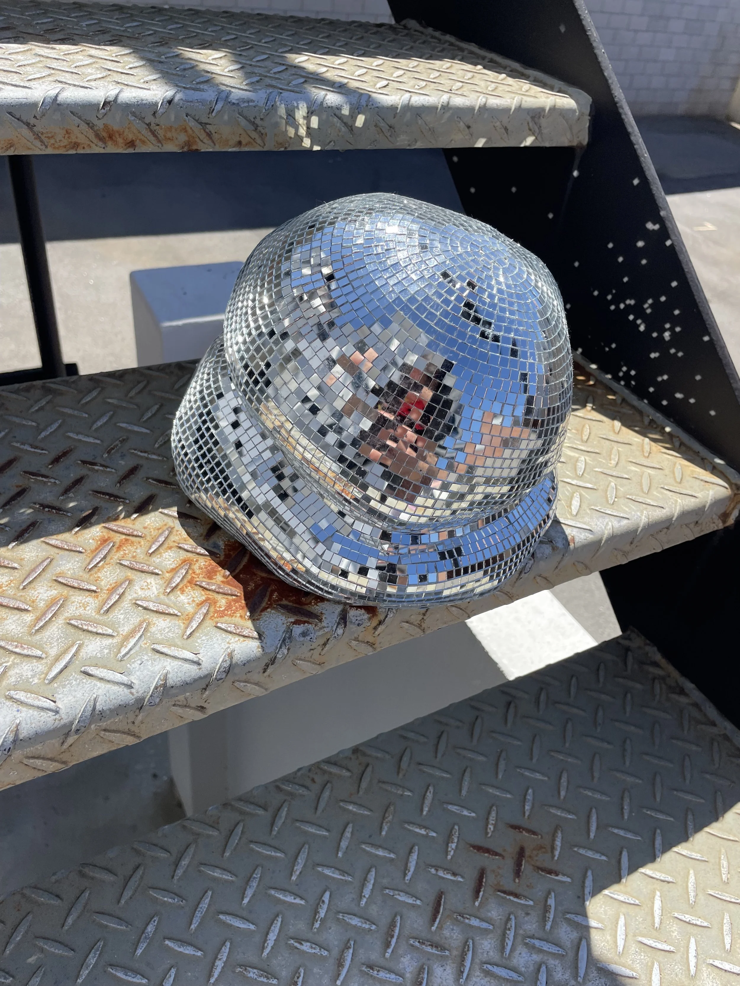 A shiny disco ball helmet resting on metal outdoor stairs with rust and dirt, reflected in its mirrored surface.