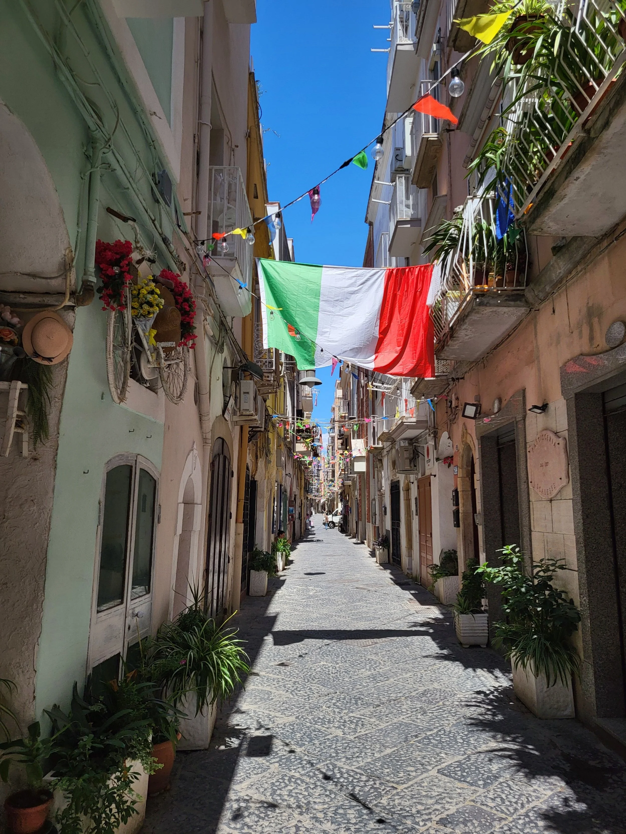 A narrow pedestrian street in a European city with colorful buildings, balconies with plants, and a large Italian flag hanging across the street. The sky is bright blue and the street is shaded with sunlight.